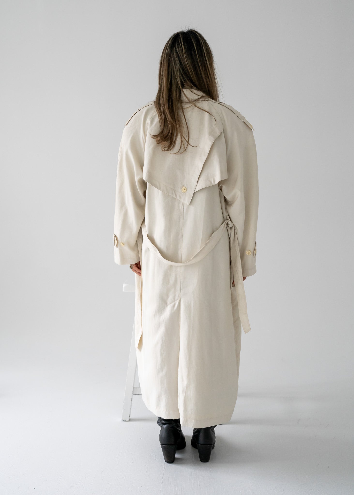 Person wearing a vintage cream-colored, double-breasted, belted, full-length trench coat on a plain background