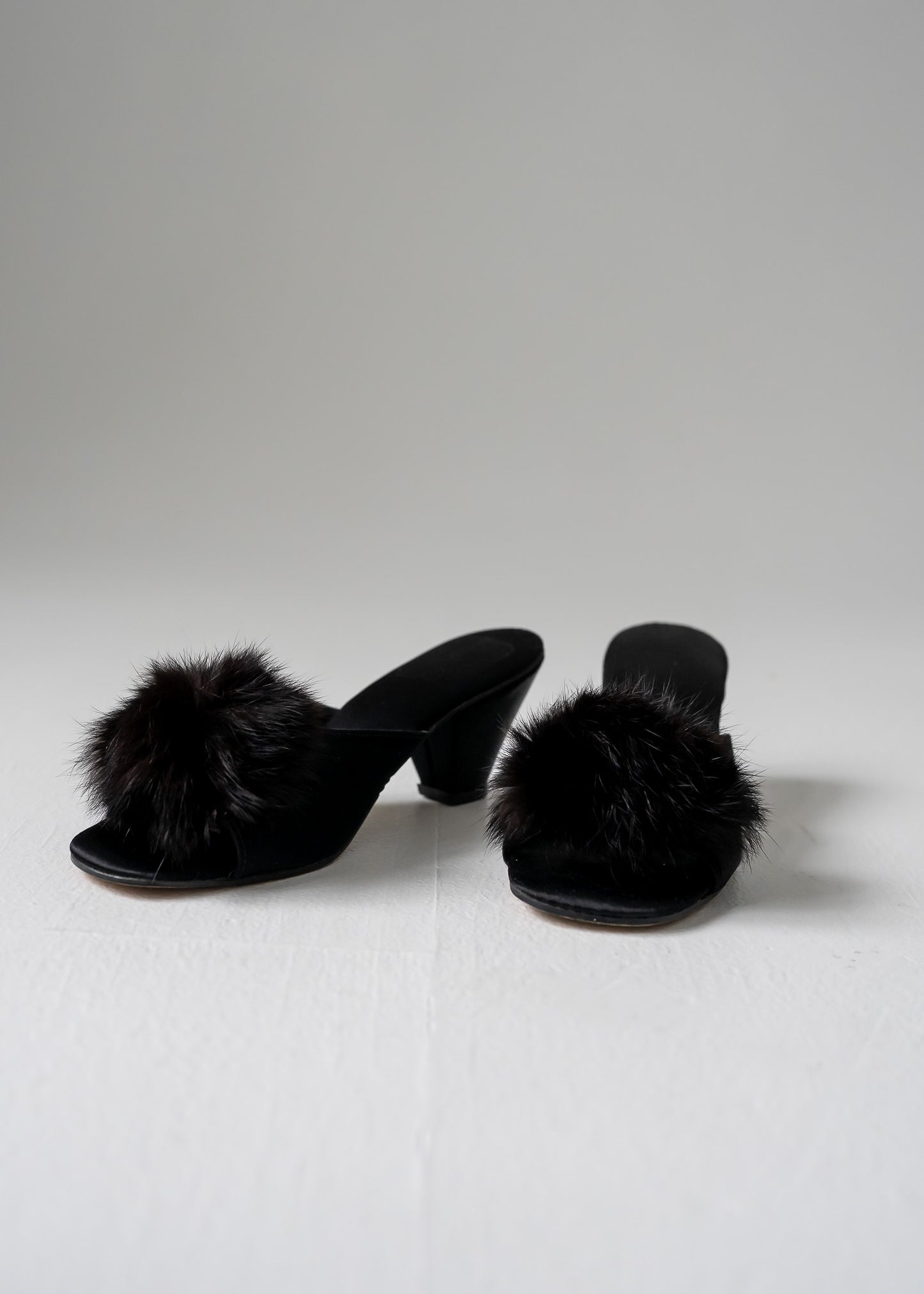 A pair of black fur pom pom kitten heels on a white background.