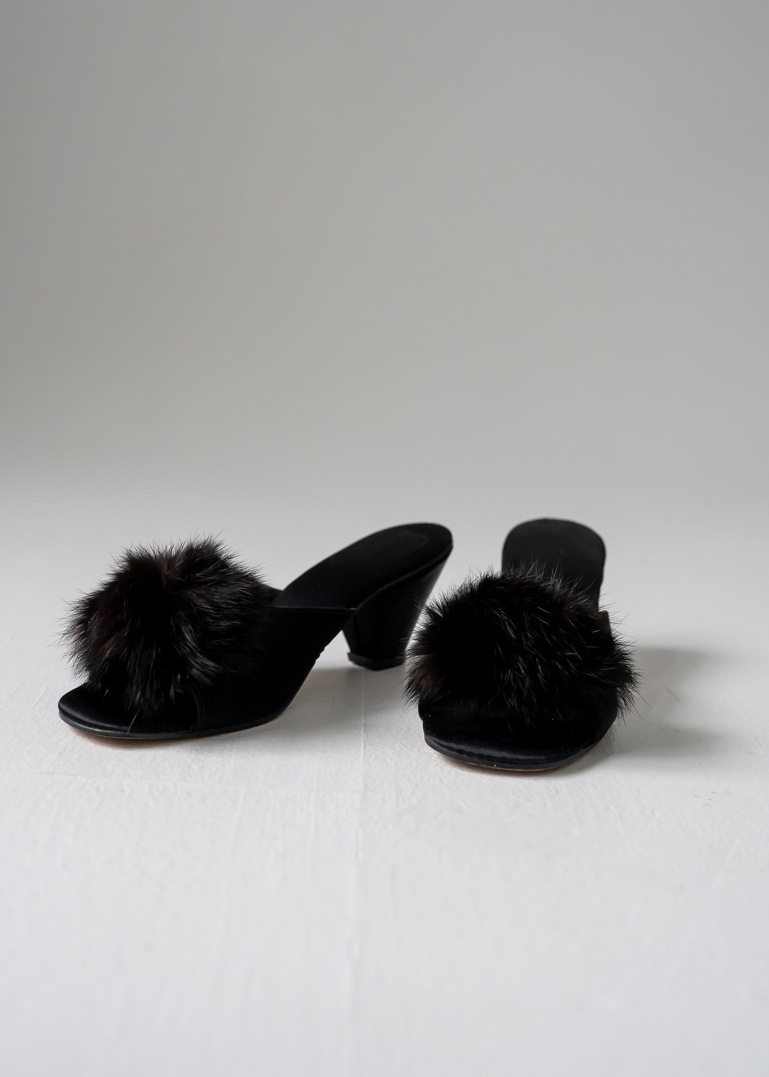 A pair of black fur pom pom kitten heels on a white background.