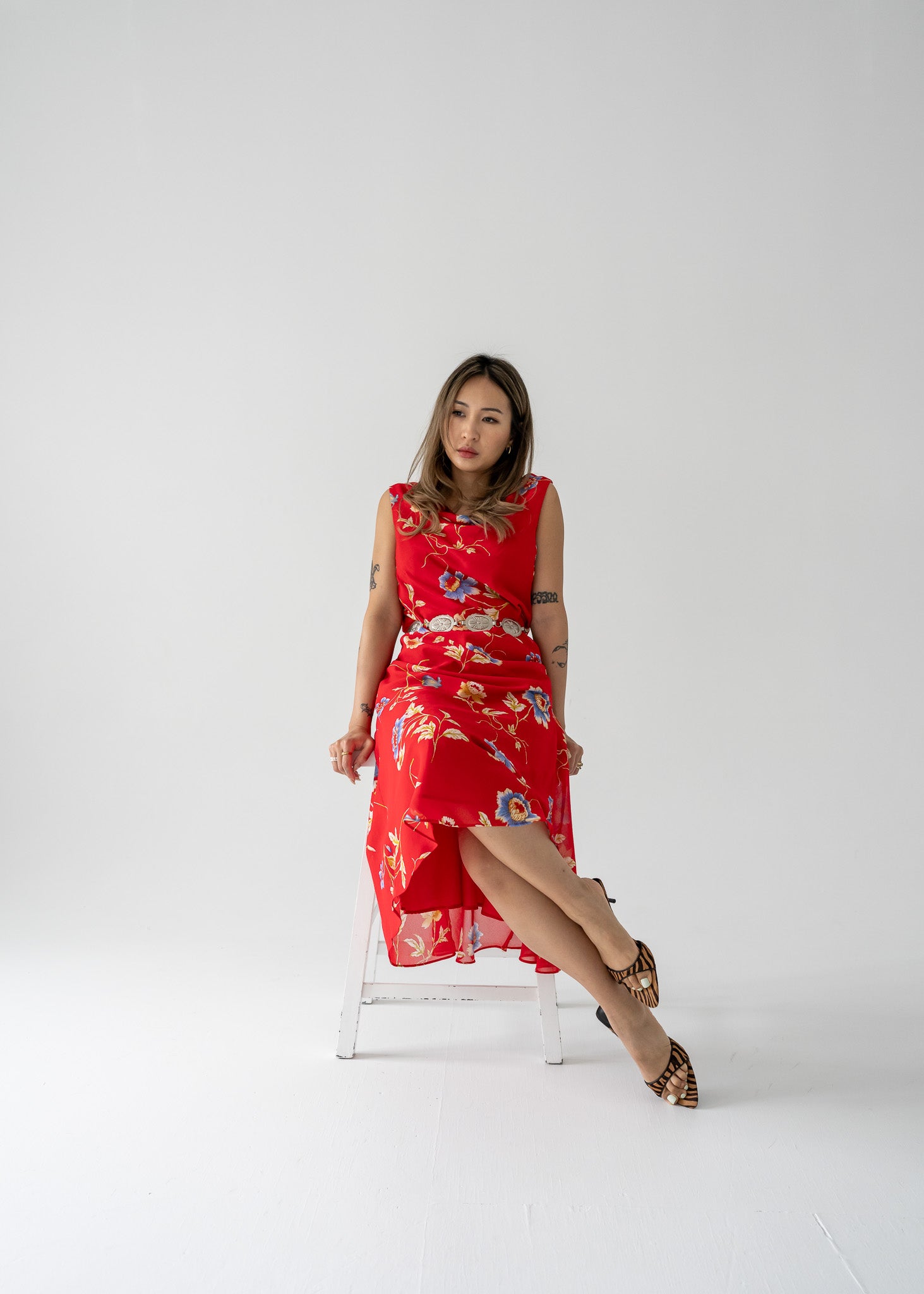A woman sitting on a white bench, wearing a bright red cowl neck bias cut floral summer dress with a left front slit.