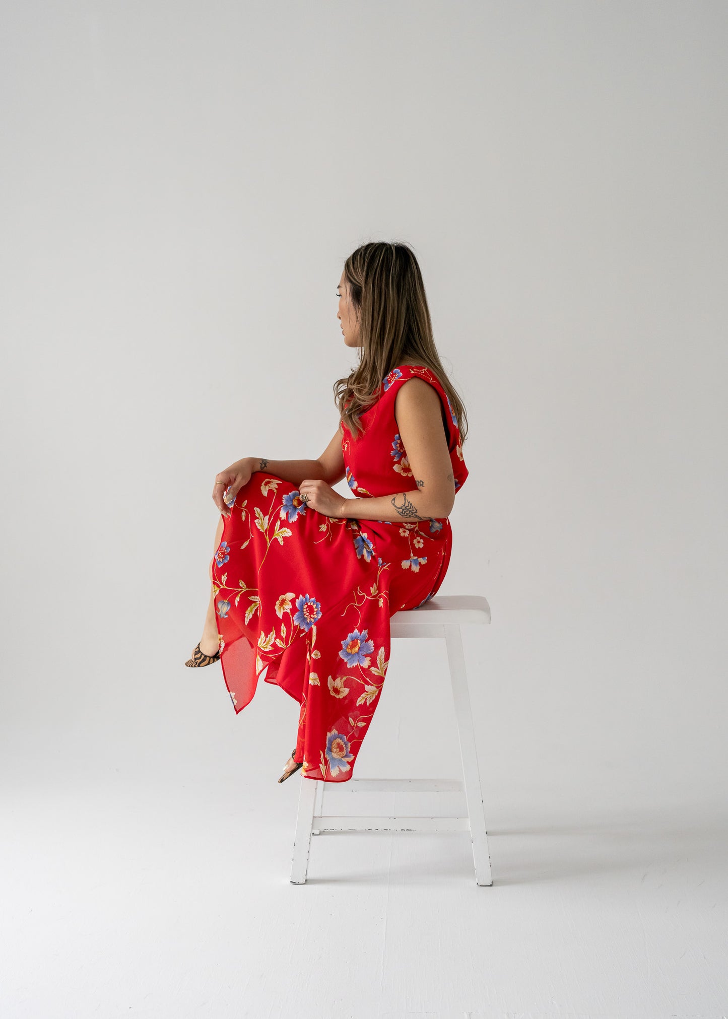 A woman sitting on a white bench, wearing a bright red cowl neck bias cut floral summer dress