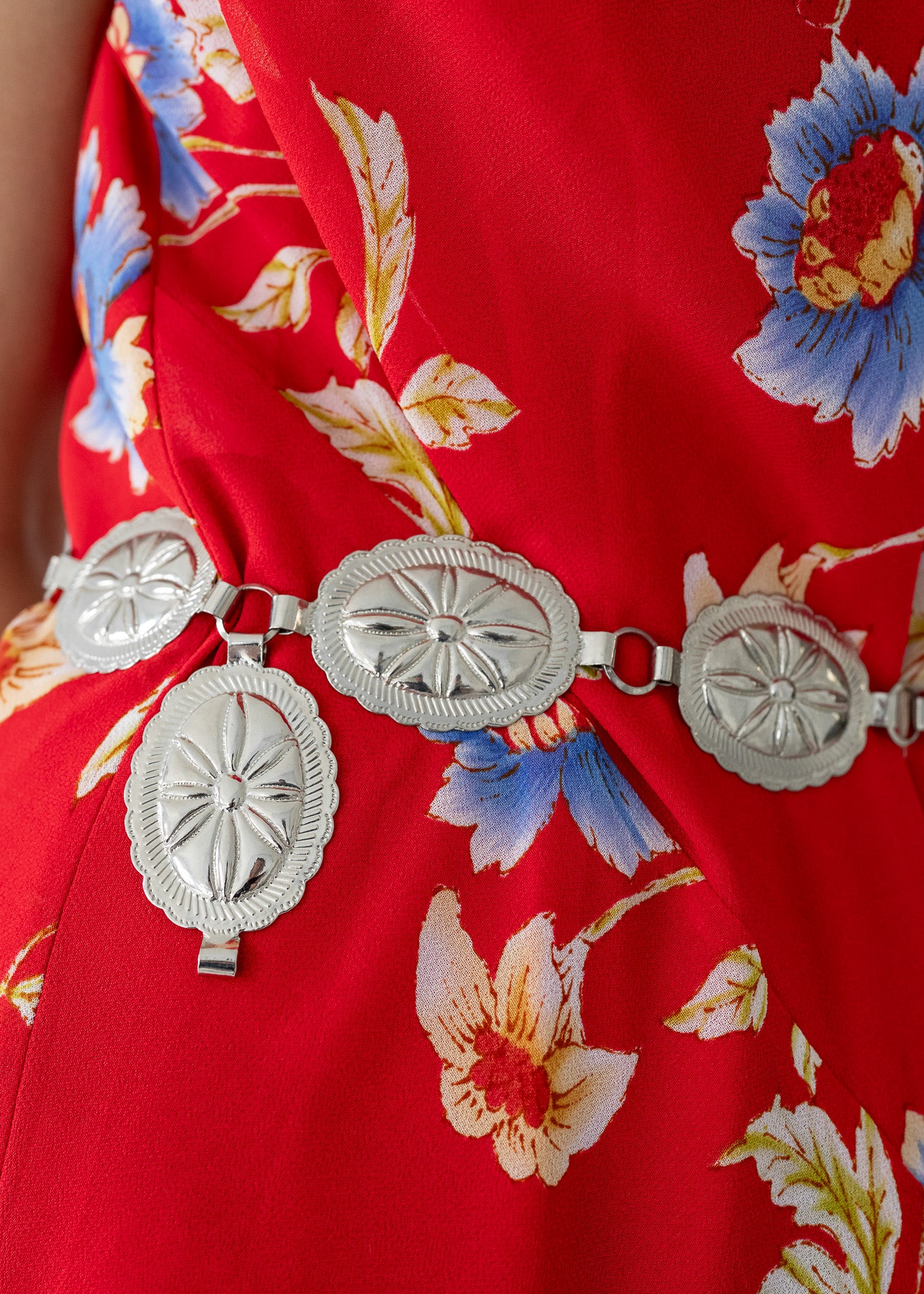 A detail shot of a red floral dress with a vintage silver tone metal stamped oval concho western style chain belt.