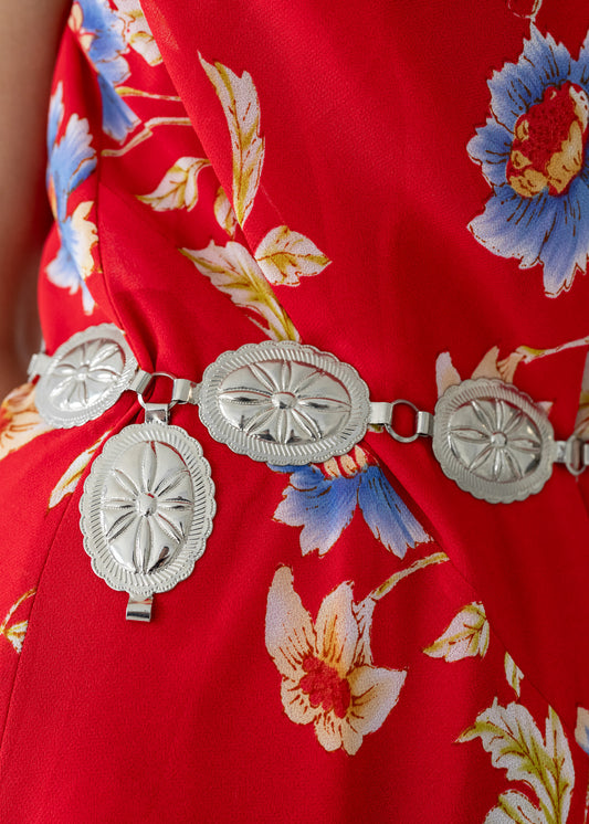 A detail shot of a red floral dress with a vintage silver tone metal stamped oval concho western style chain belt.