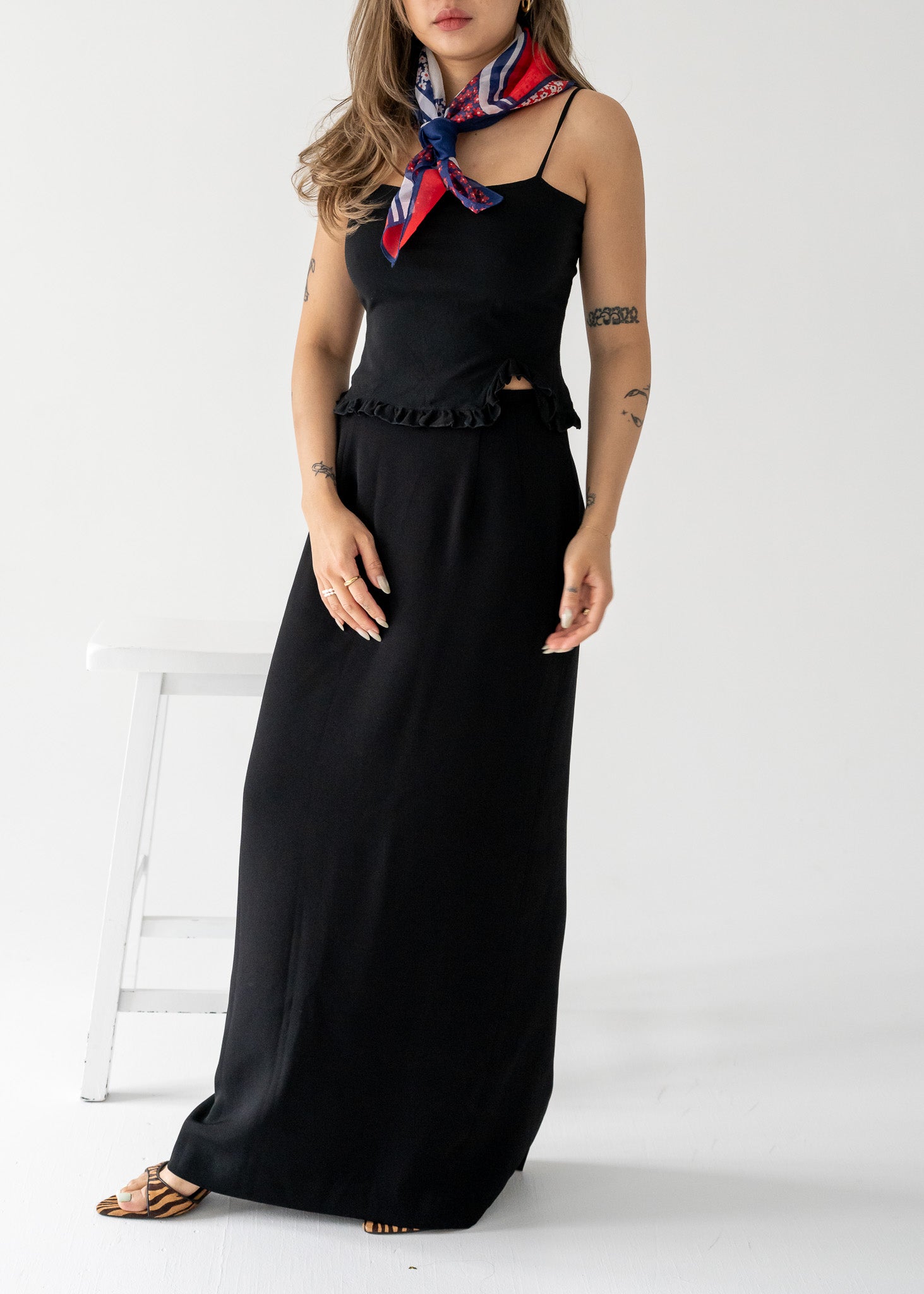 Woman wearing a vintage black tank top with a ruffle hem and black column skirt with a red and blue scarf around her neck, standing against a white background.