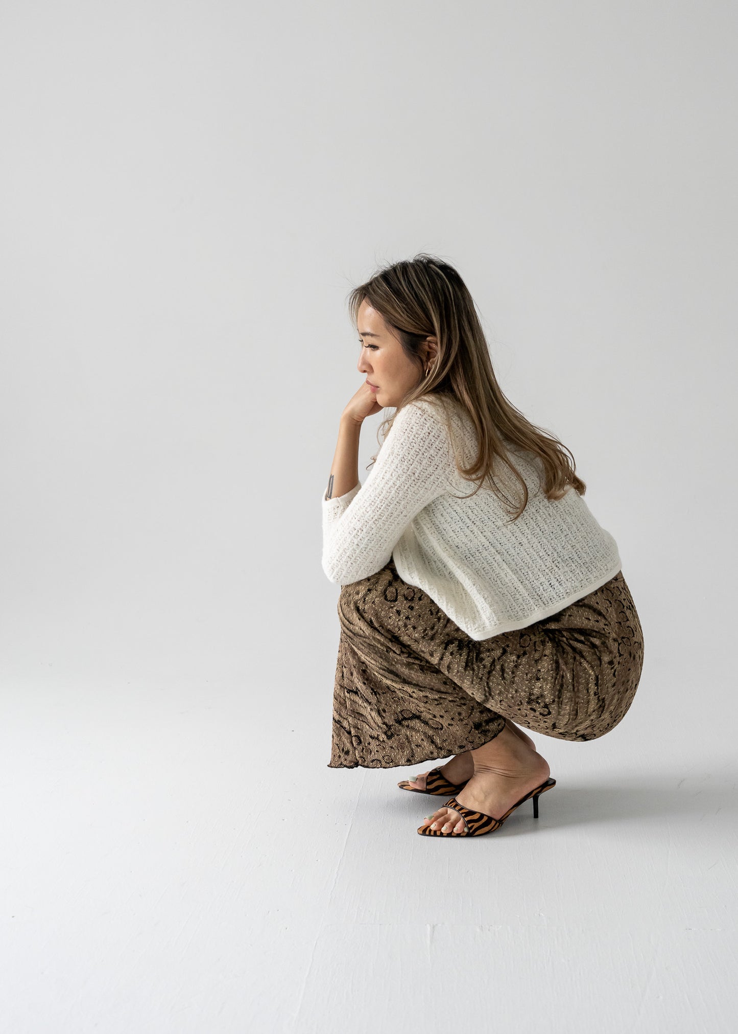 Woman in white knitted top and vintage long maxi skirt with a bronze and black abstract animal print squatting on a plain background