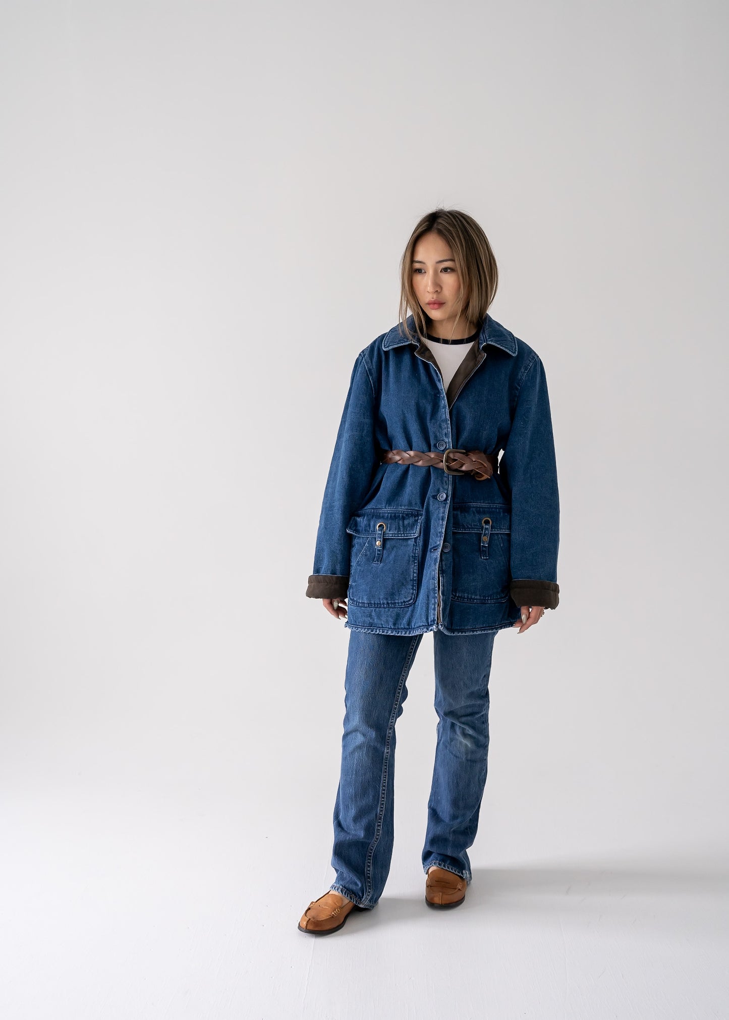 Person wearing a blue denim jacket and jeans with a braided leather belt on a white background