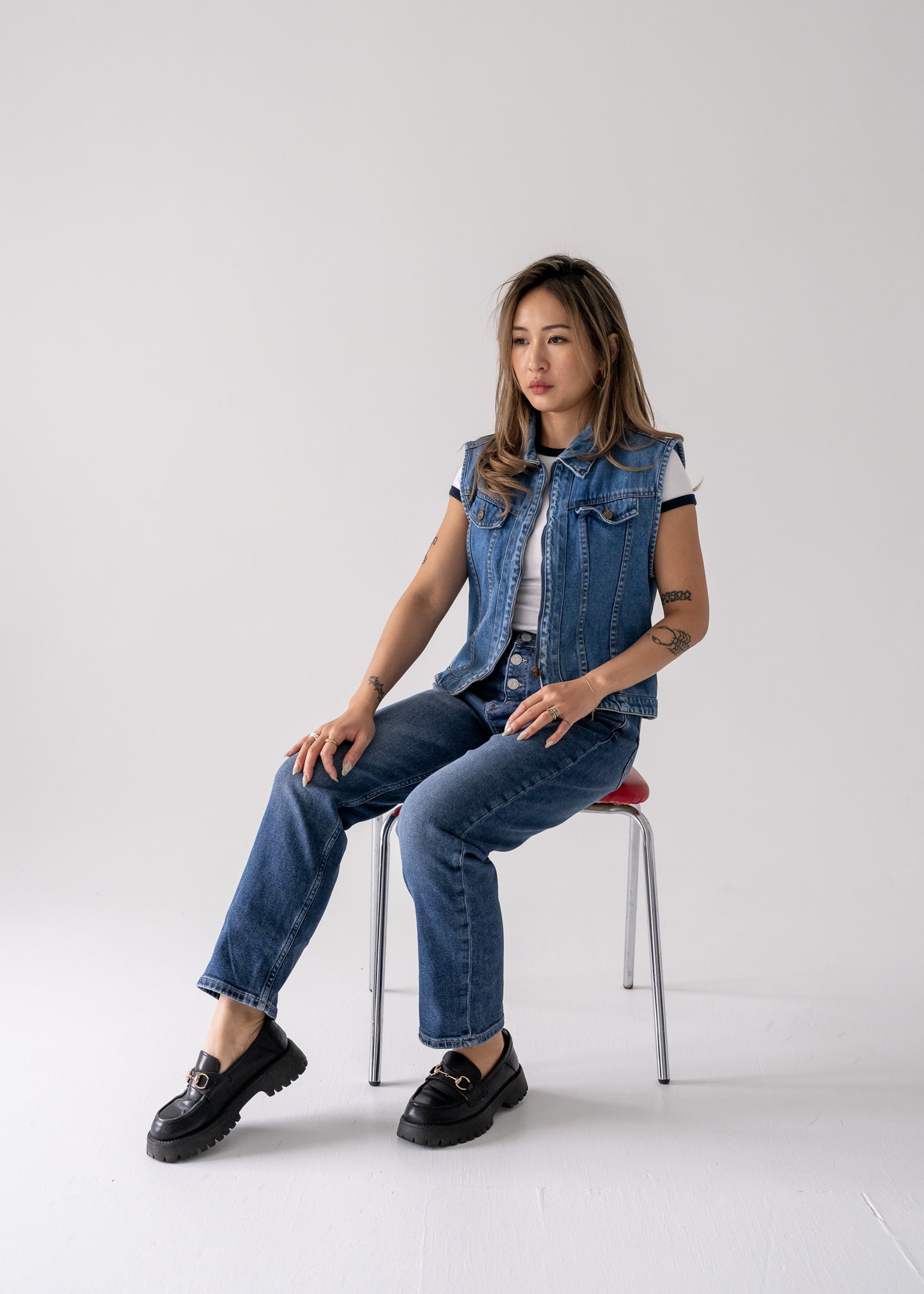 A person seated on a chair wearing a vintage medium wash blue denim vest with a collared zip front, blue jeans, and black shoes.