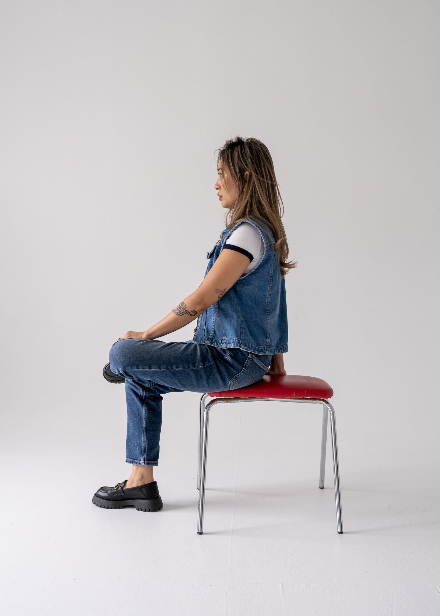 Person wearing a vintage medium wash blue denim vest with a collared zip front and blue jeans sitting on a red chair against a plain background