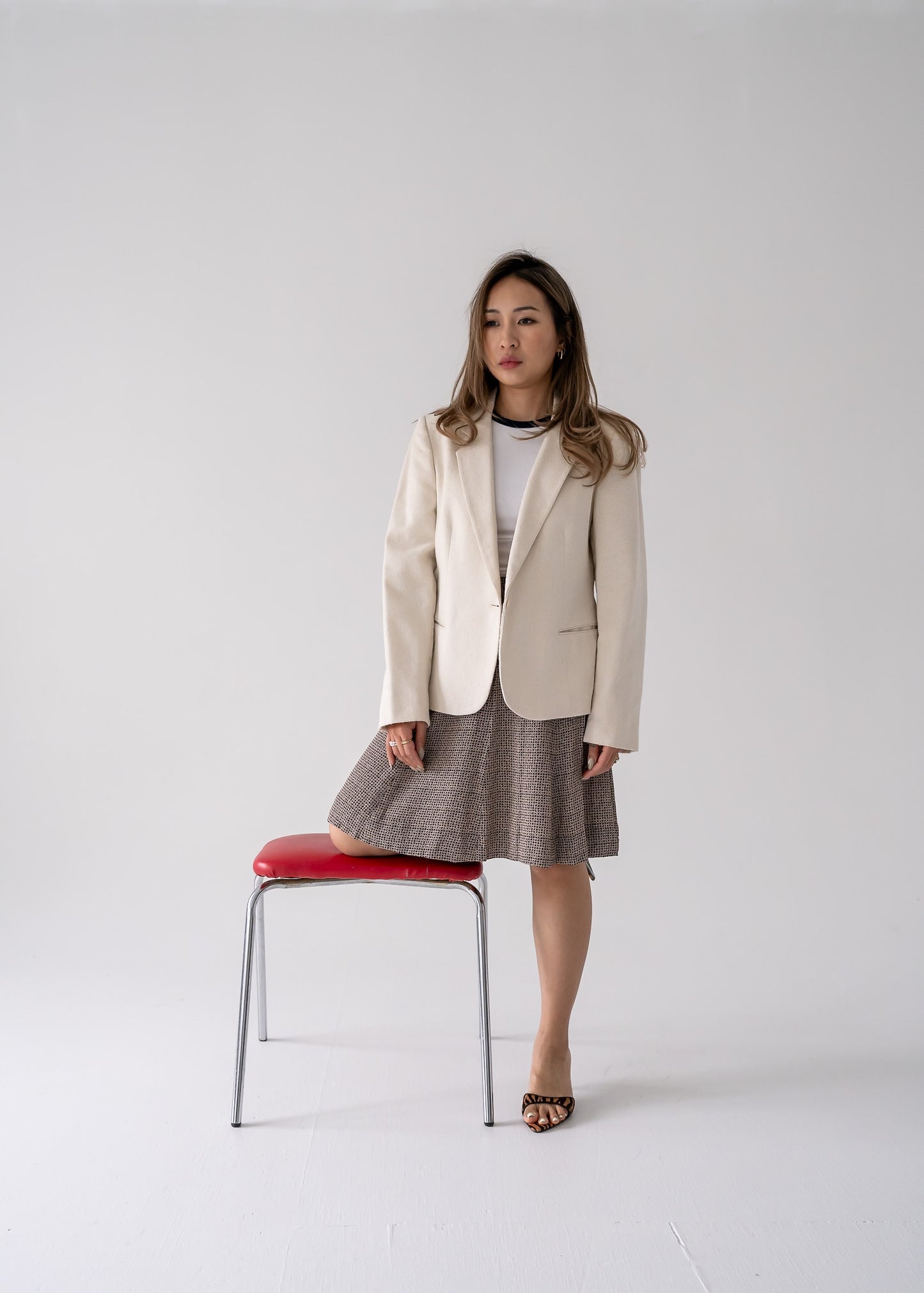 A person wearing a vintage cream-colored, one-button blazer with pockets, paired with a patterned skirt and standing next to a red stool.