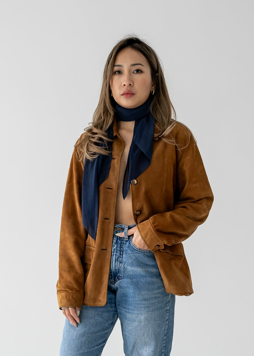 A person wearing a navy blue micro pleat crinkle scarf with a brown suede jacket and blue jeans.