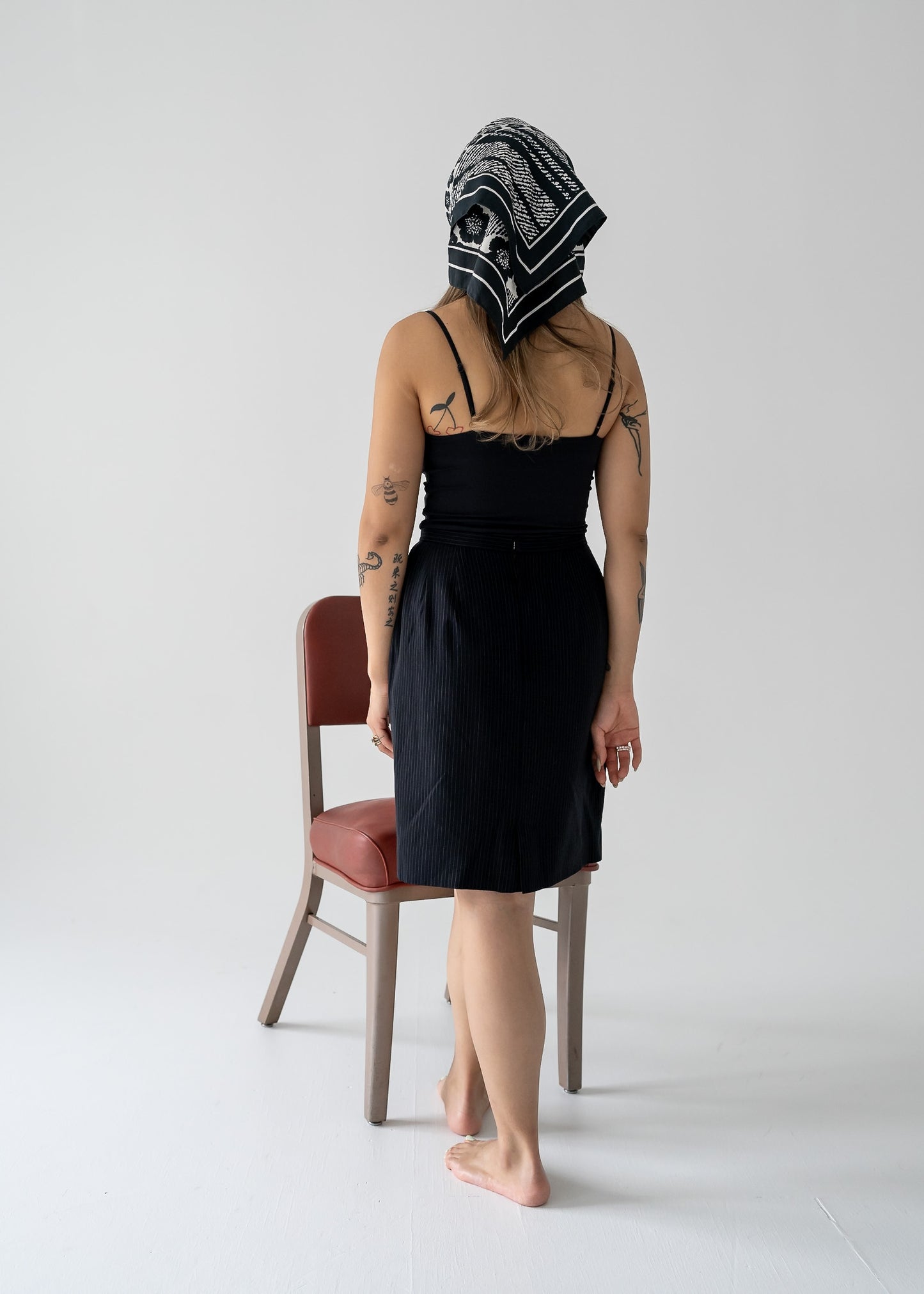 Person in a vintage fitted A-line mini skirt in black and white stripes and black tank top with a headscarf, standing by a chair on a white background.