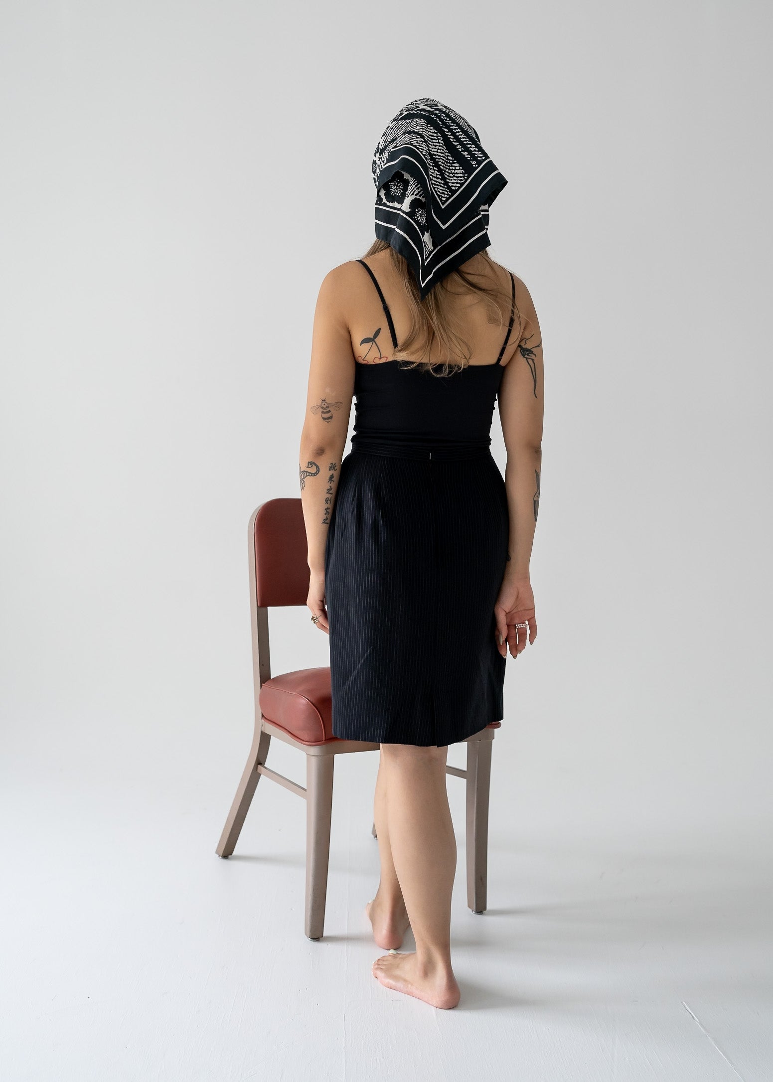 Person in a vintage fitted A-line mini skirt in black and white stripes and black tank top with a headscarf, standing by a chair on a white background.