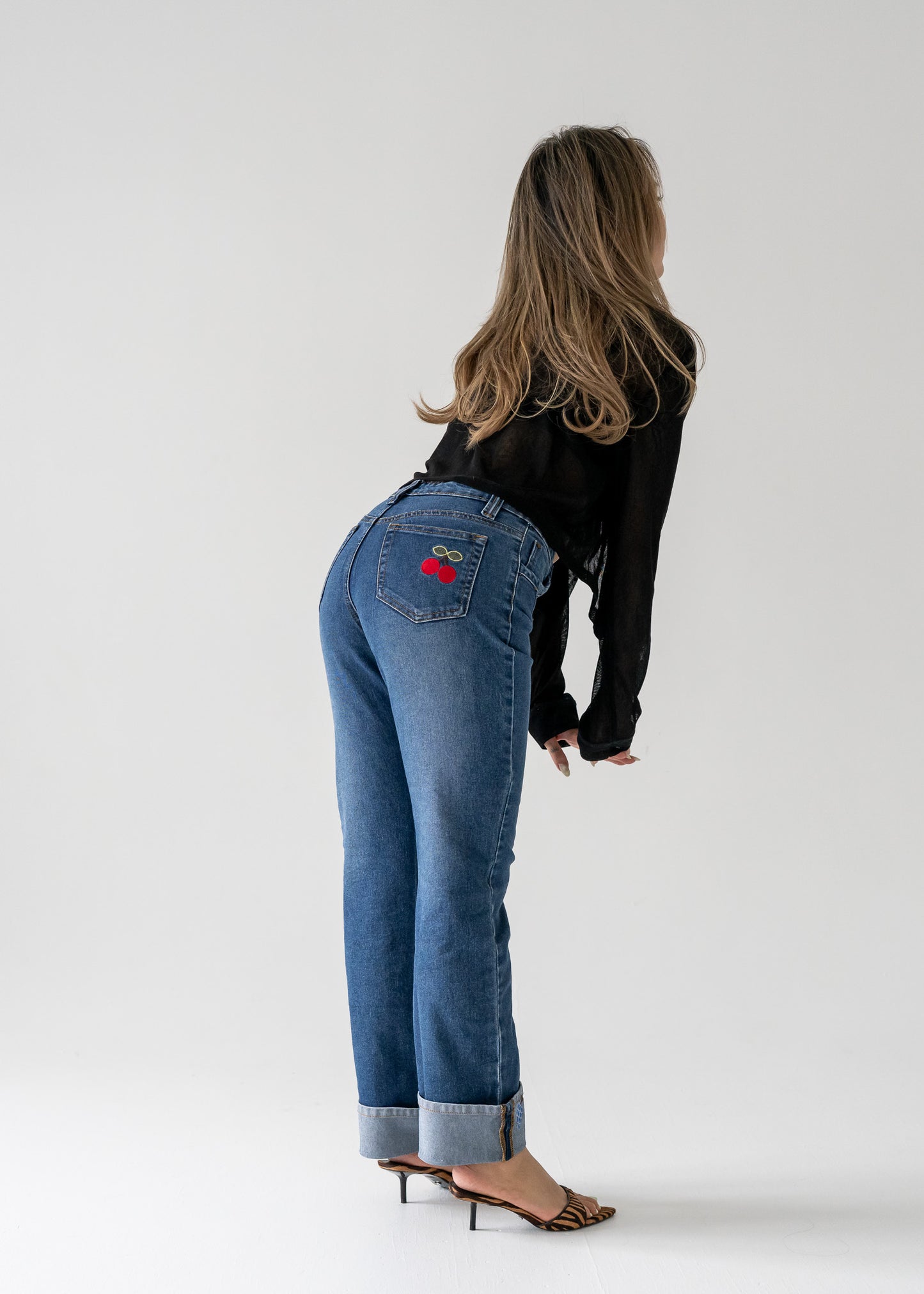 Person wearing vintage medium wash blue jeans with cuffed hem with a red cherry design on the back pocket against a white background
