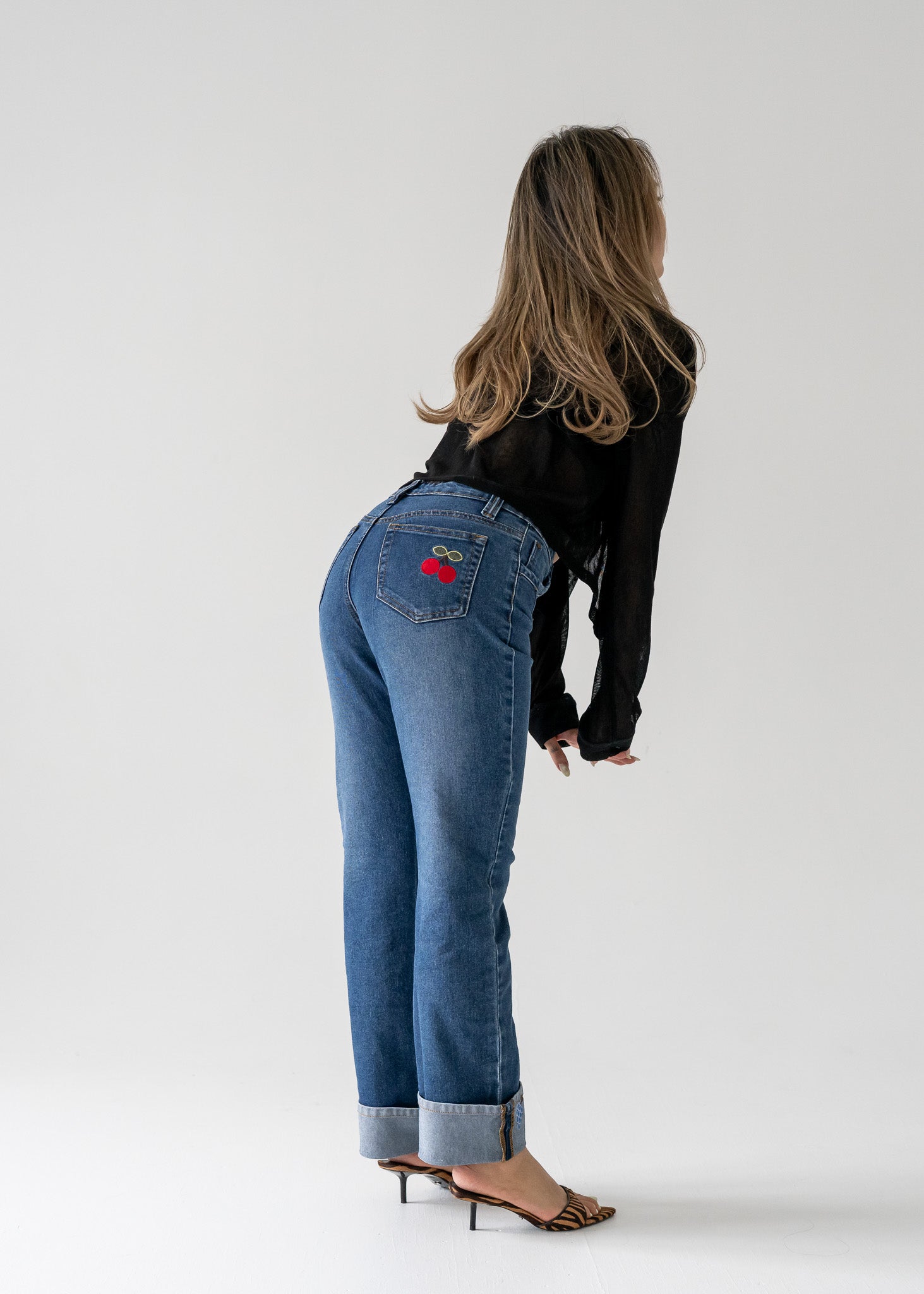 Person wearing vintage medium wash blue jeans with cuffed hem with a red cherry design on the back pocket against a white background