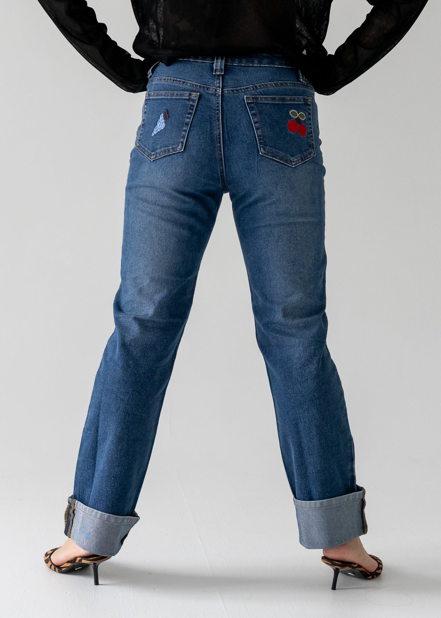 Person wearing vintage medium wash blue jeans with cuffed hem with a red cherry design on the back pocket against a white background