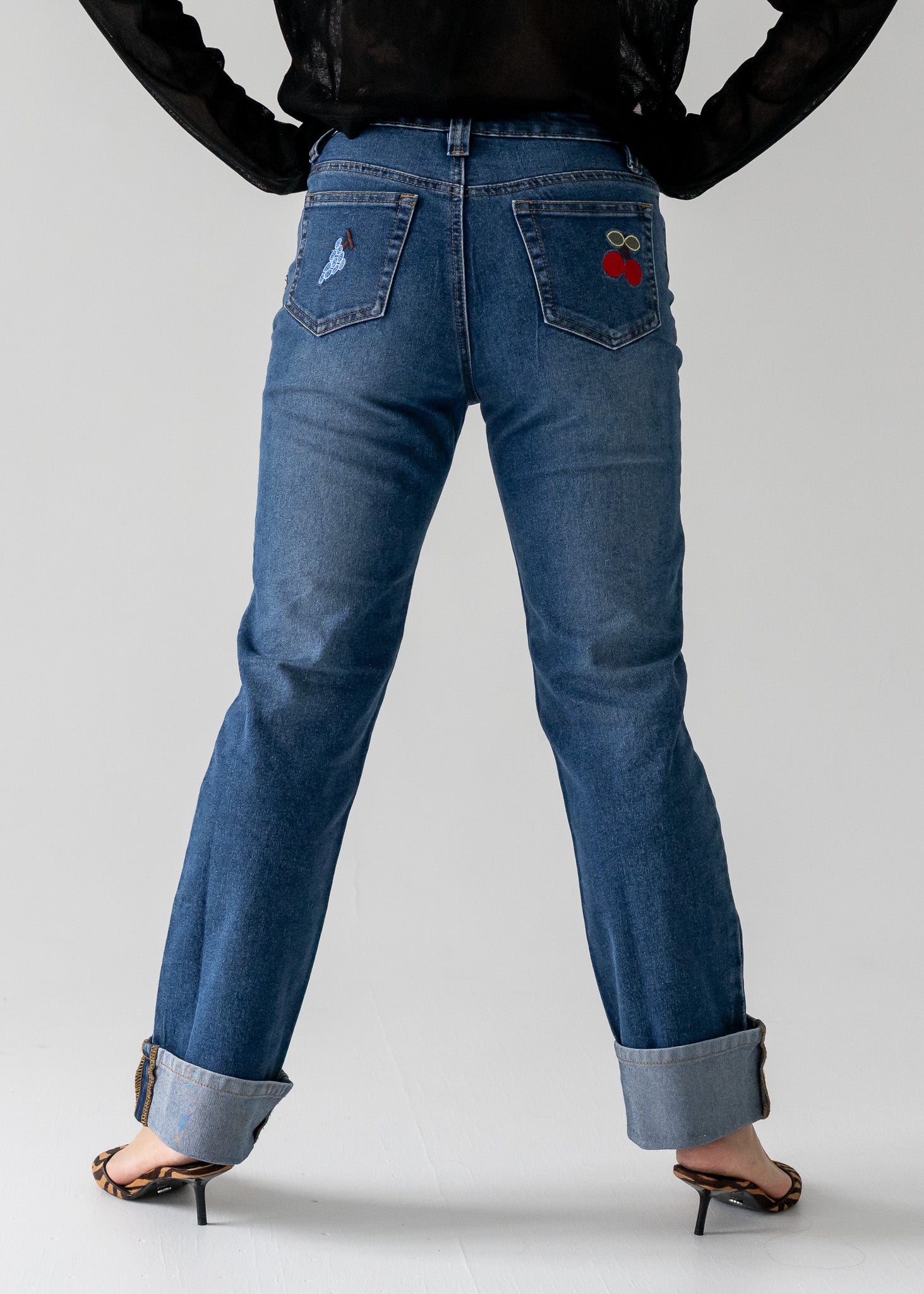 Person wearing vintage medium wash blue jeans with cuffed hem with a red cherry design on the back pocket against a white background