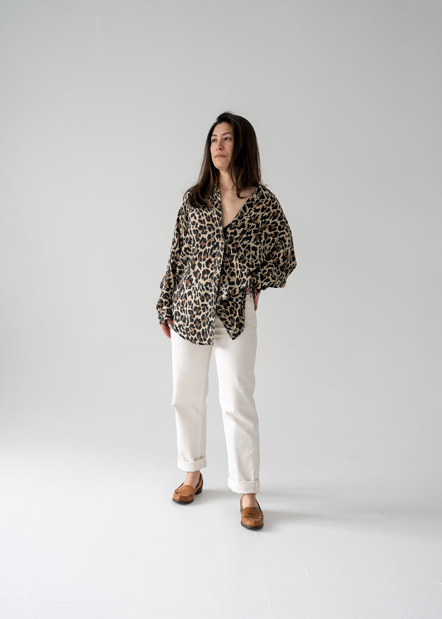 A person standing against a white background, wearing a vintage silk leopard animal print long sleeve button up blouse with a classic collar, paired with light-colored pants and tan shoes.