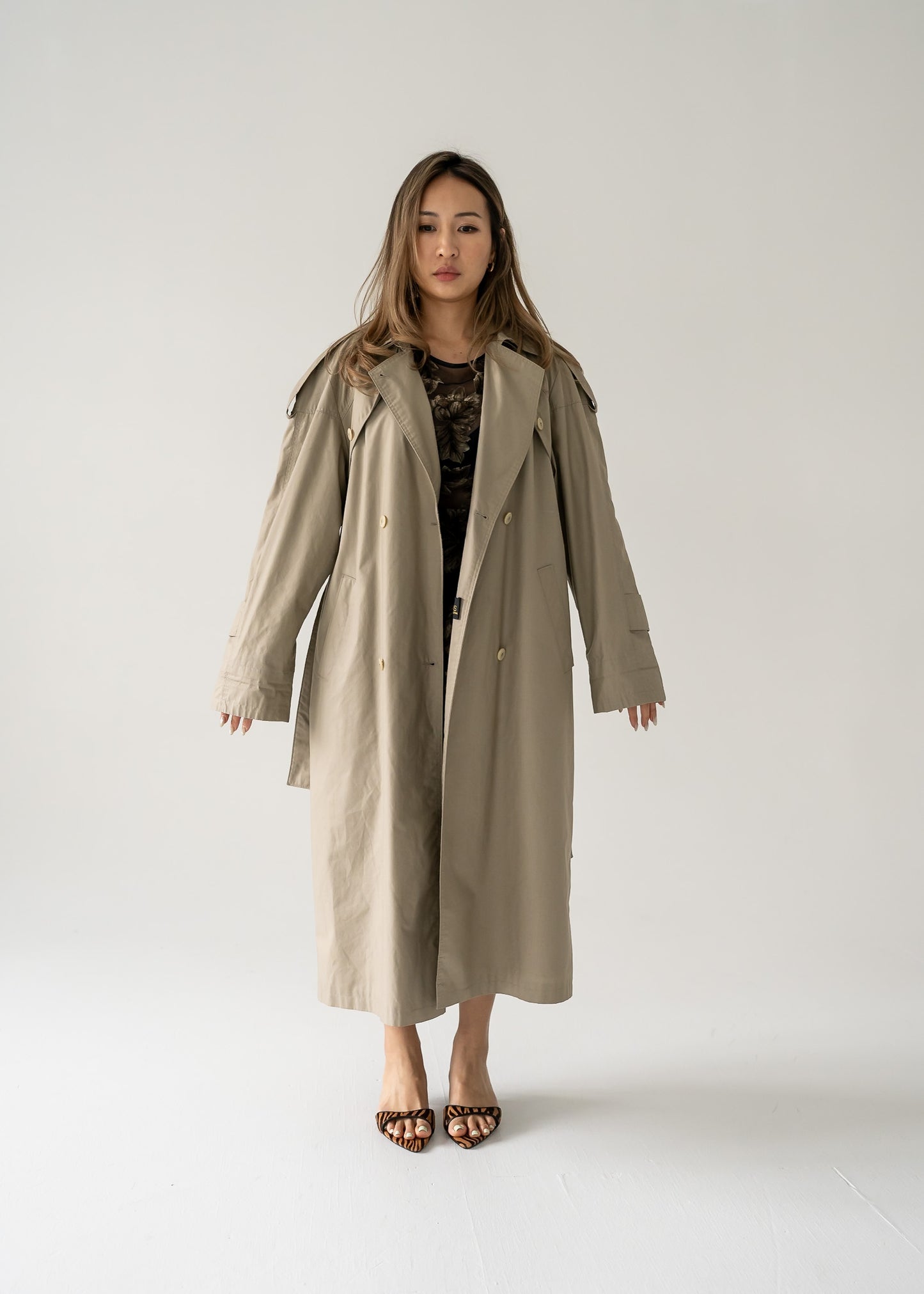 A person standing in a studio wearing a classic khaki long button-up trench coat with a double-breasted button closure, a classic collar, and a belted waist.