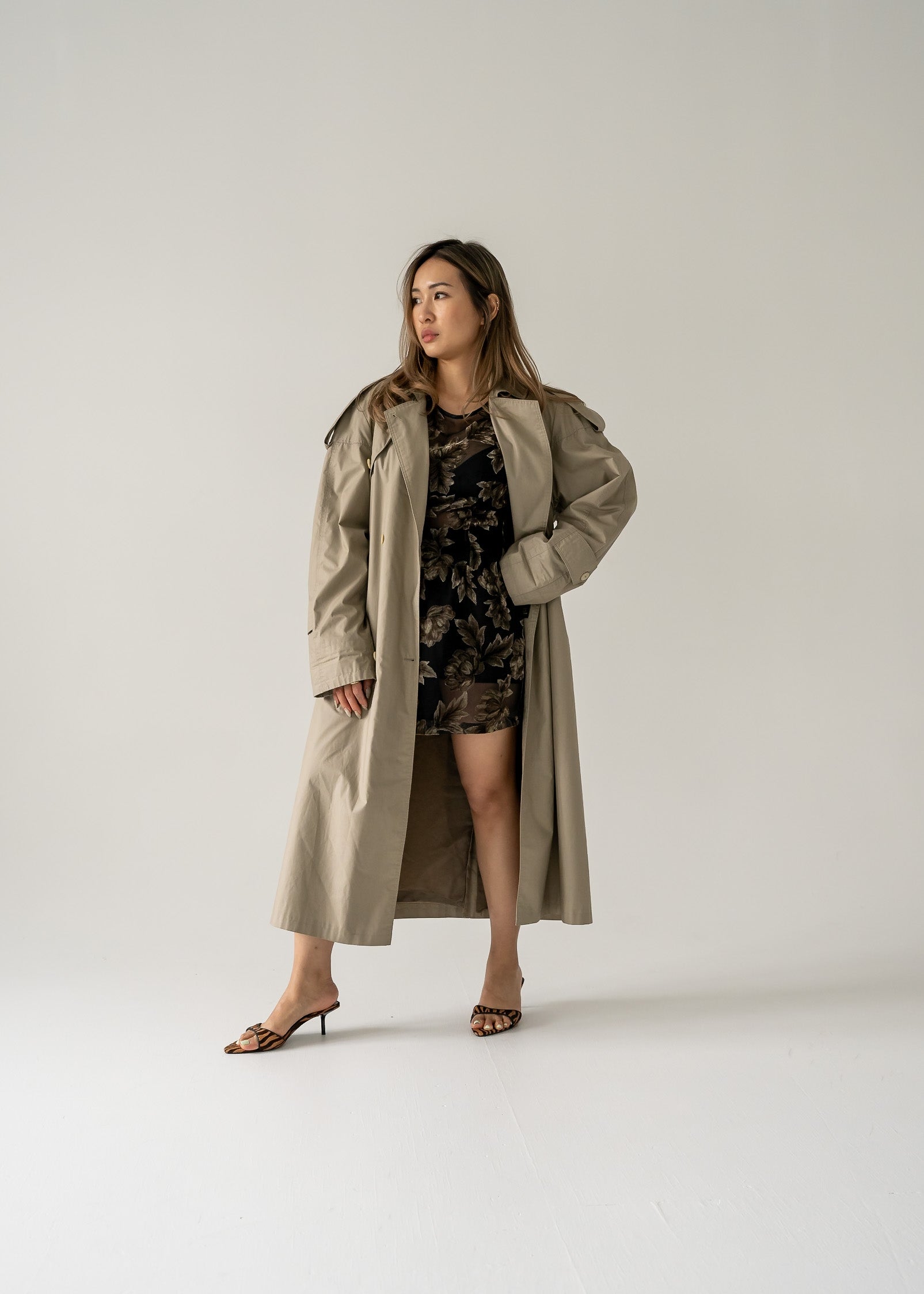 Woman wearing a beige trench coat over a vintage dark green and black floral velvet burnout sheer sleeveless mini dress on a plain background