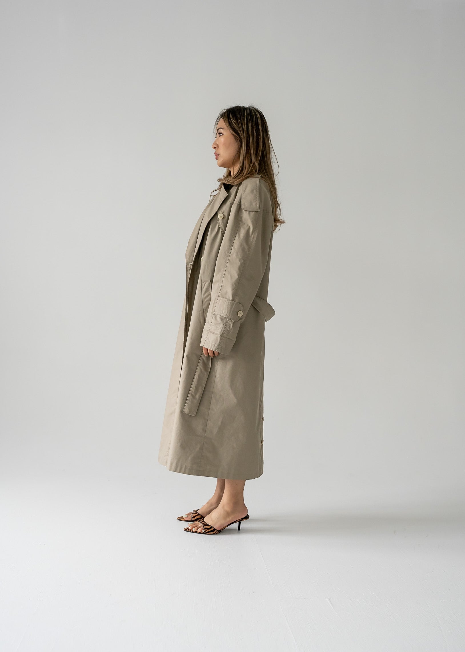 A person standing in a studio wearing a classic vintage khaki long button-up trench coat with a double-breasted button closure, a classic collar, and a belted waist.