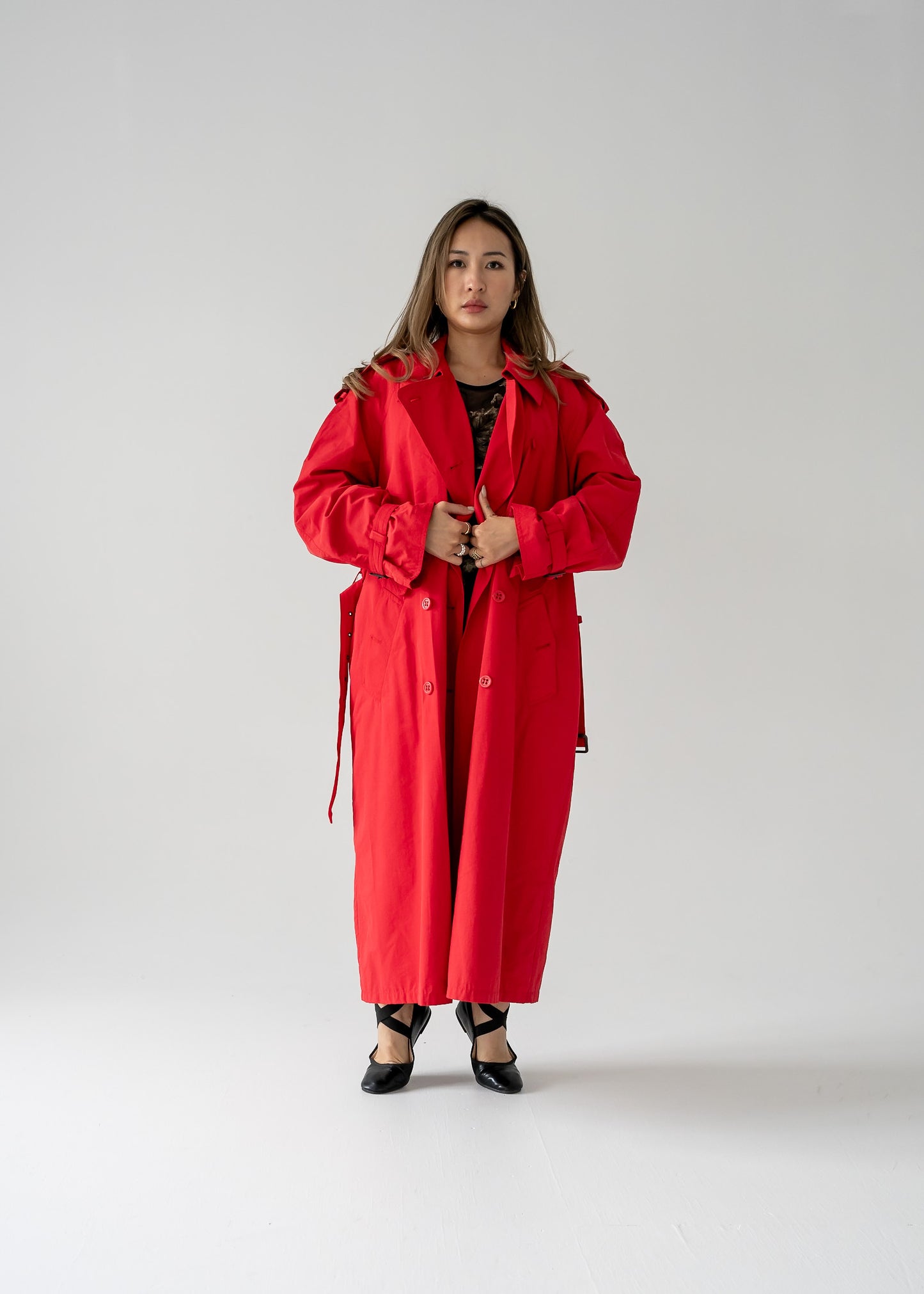 A person standing in a studio setting, wearing a vintage cherry red double breasted button up belted full length trench coat.