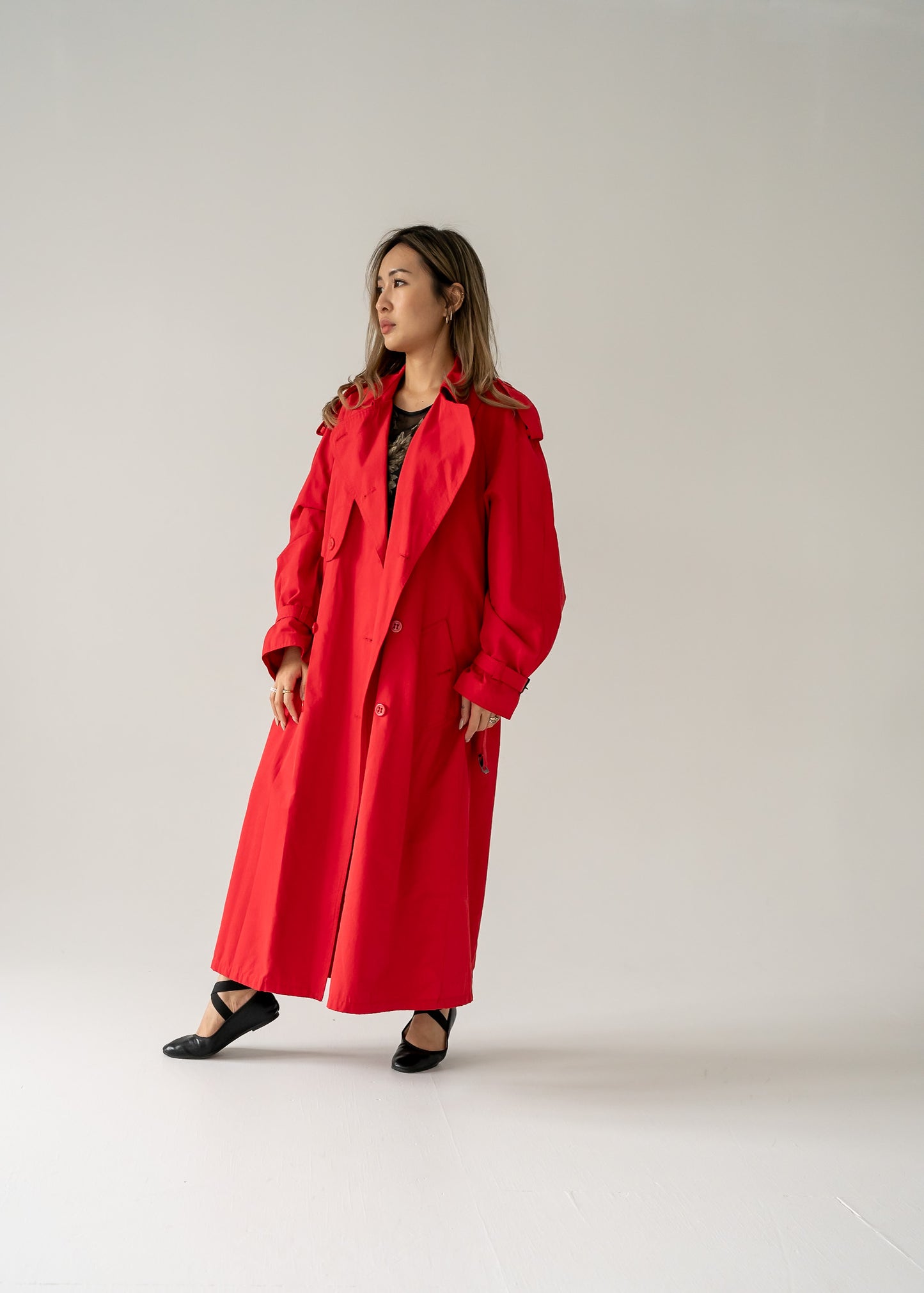 A person standing in a studio setting, wearing a vintage cherry red double breasted button up belted full length trench coat.
