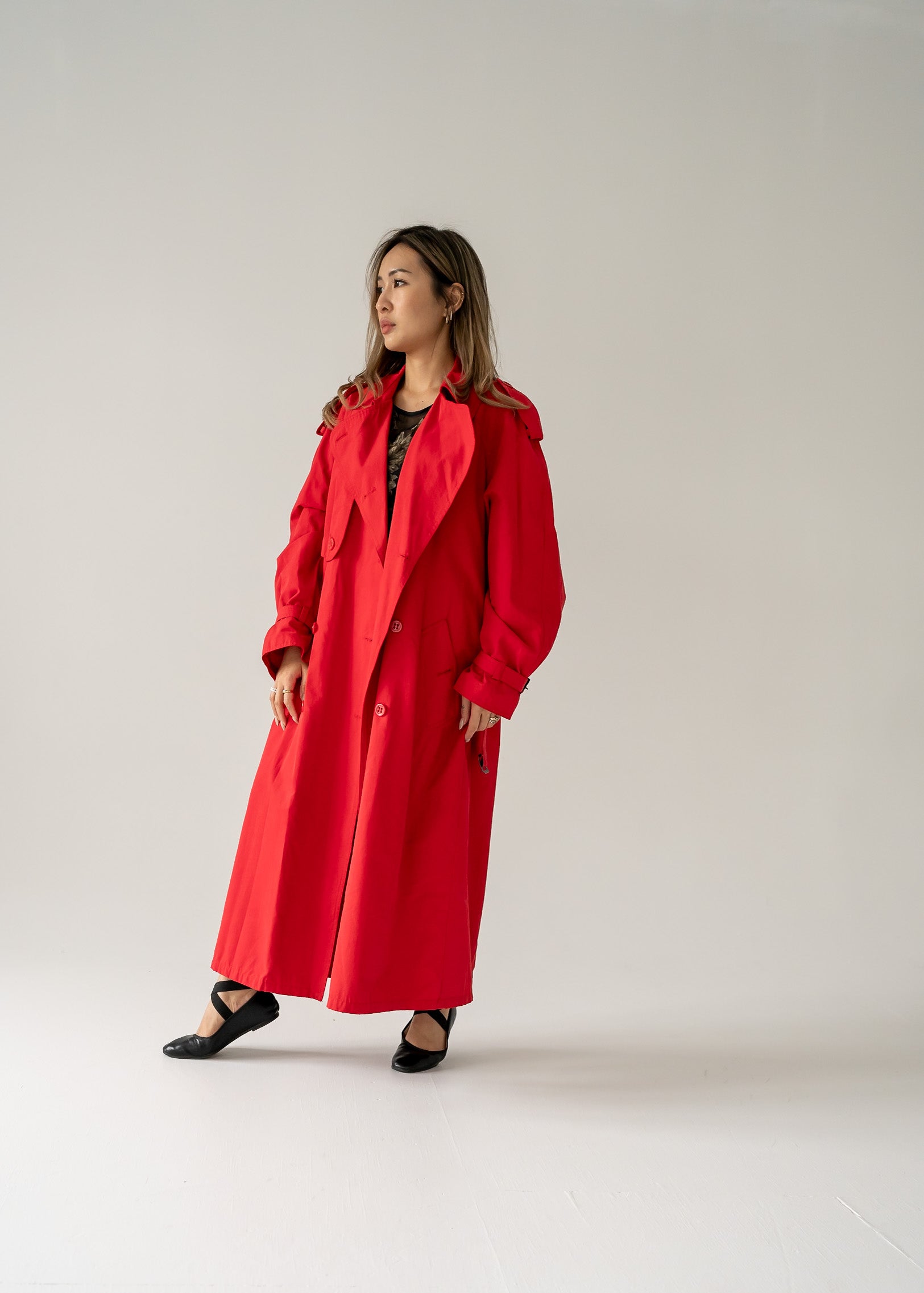 A person standing in a studio setting, wearing a vintage cherry red double breasted button up belted full length trench coat.