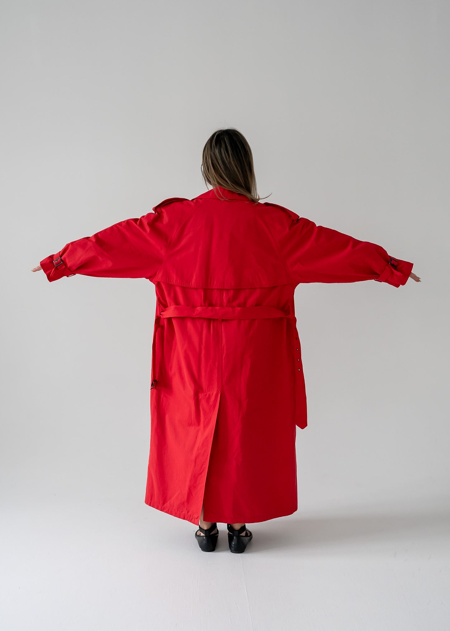 A person standing in a studio setting, wearing a vintage cherry red double breasted button up belted full length trench coat.