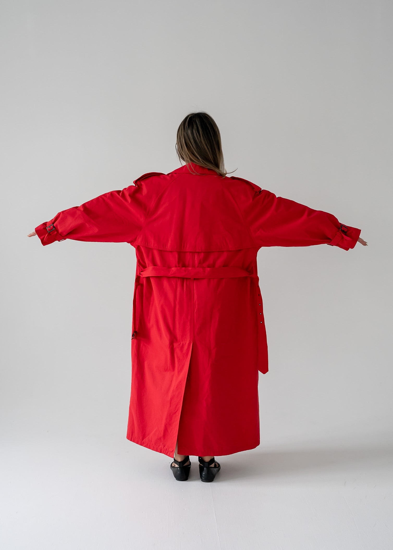 A person standing in a studio setting, wearing a vintage cherry red double breasted button up belted full length trench coat.