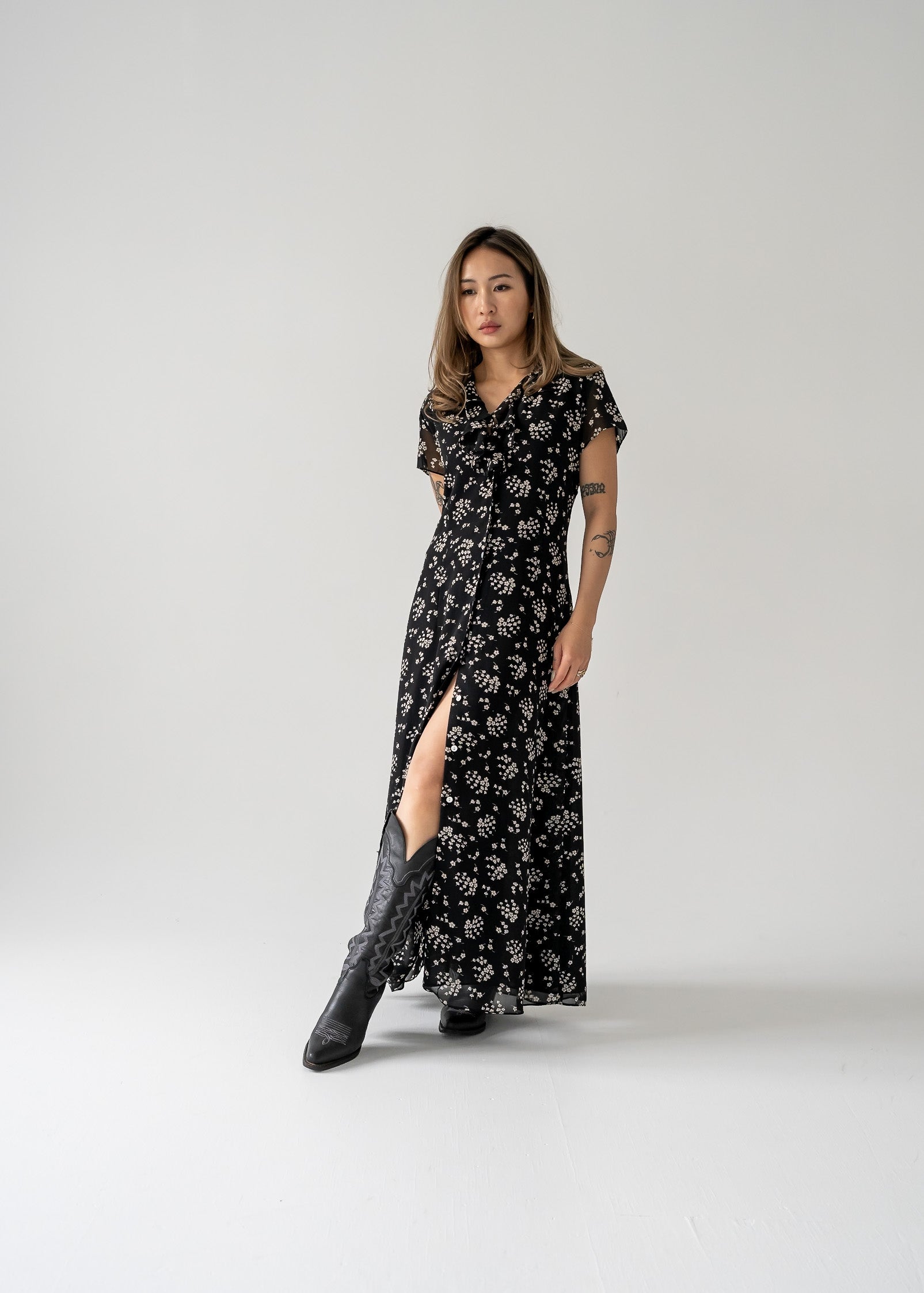 A model wearing a vintage black and cream small print floral ruffle neck button up cap sleeve maxi dress with sheer black lining, standing against a white background.