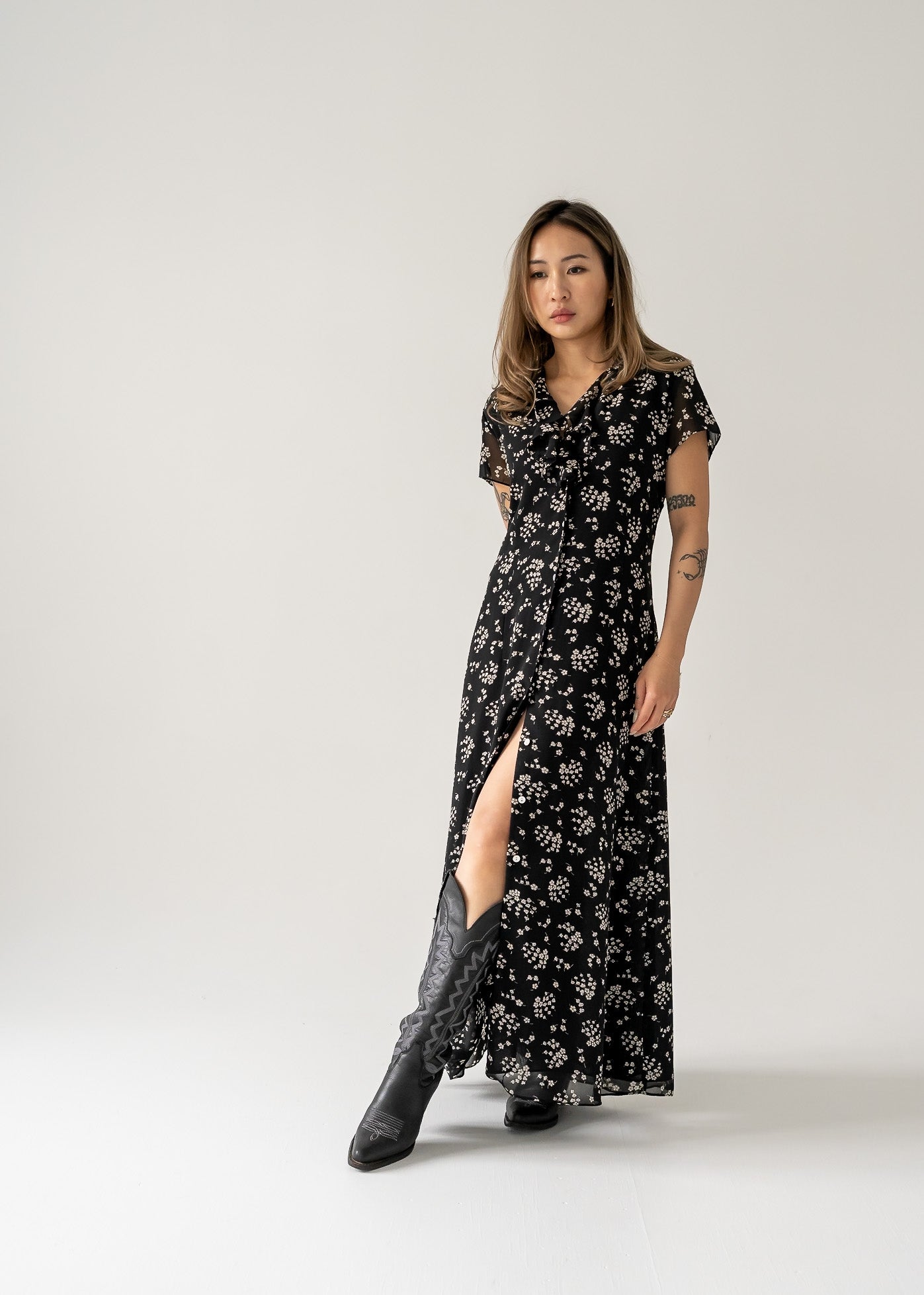 Woman in a vintage black and cream small print floral ruffle neck button up cap sleeve maxi dress with a slit, walking on a plain background.