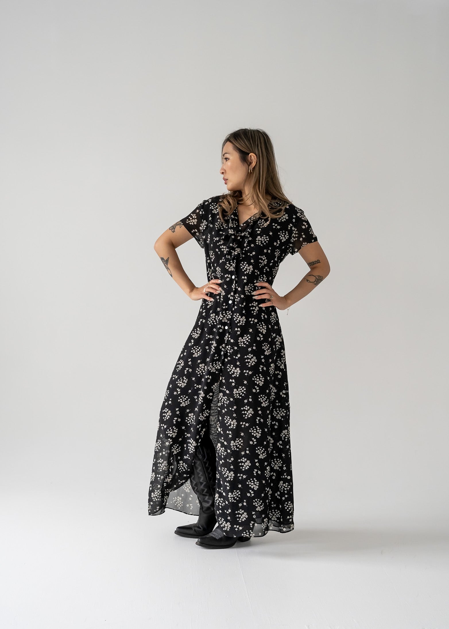 Woman wearing a vintage black and cream small print floral ruffle neck button up cap sleeve maxi dress on a white background
