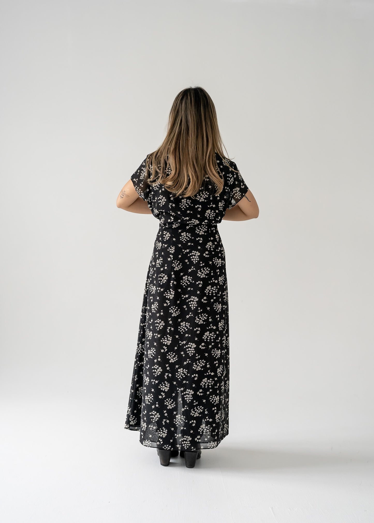 Woman wearing a vintage black and cream small print floral ruffle neck button up cap sleeve maxi dress on a white background