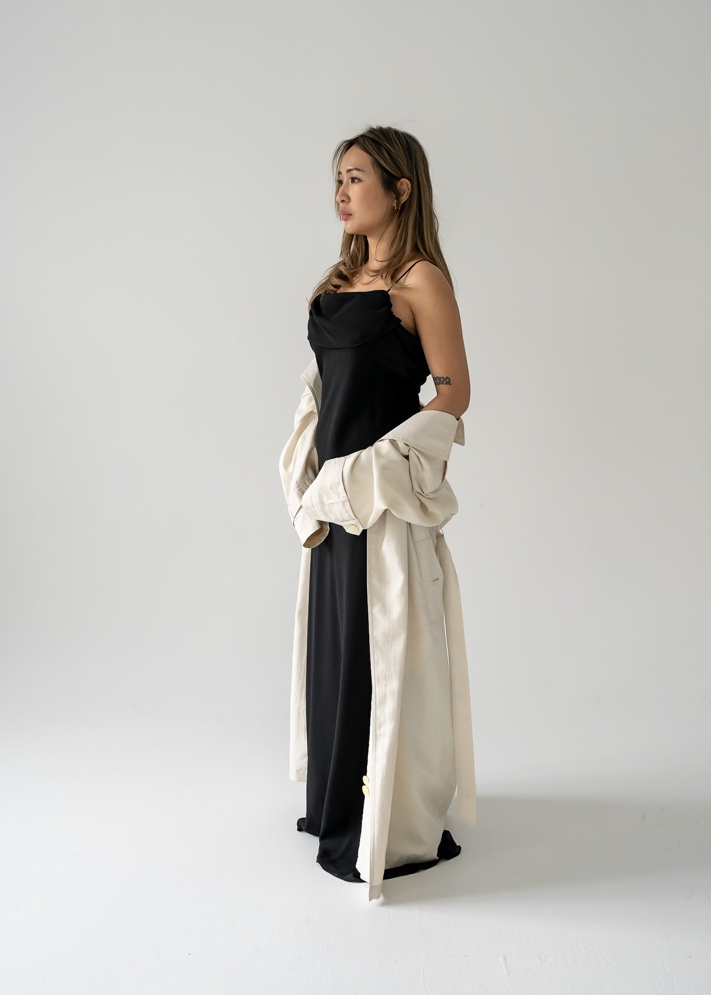 A woman standing in a studio setting, wearing a black floor-length crepe evening dress with a triple draped cowl collar and thin straps.