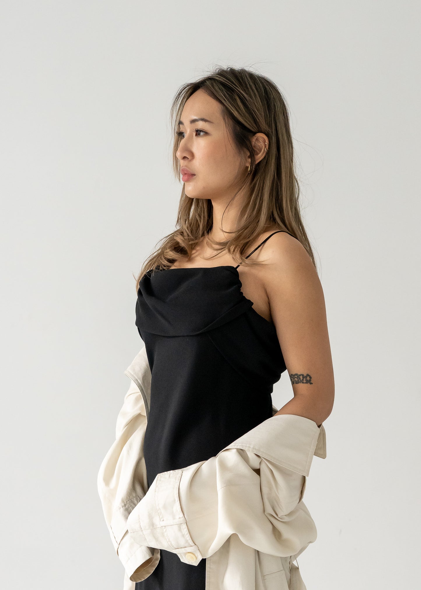 Woman wearing a vintage black crepe sleeveless dress and holding a beige jacket against a plain background
