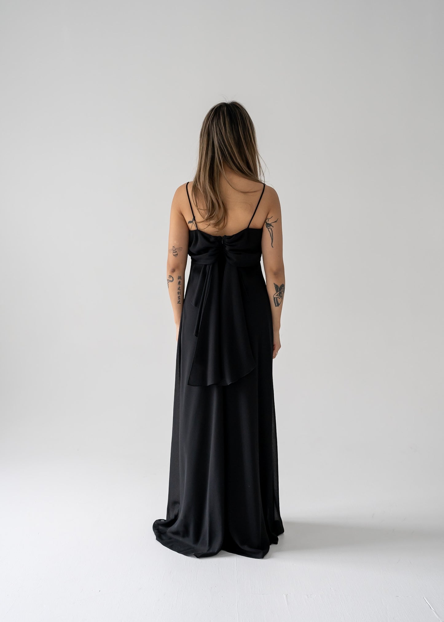 A woman standing in a studio setting, wearing a black floor-length crepe evening dress with a triple draped cowl collar and thin straps.