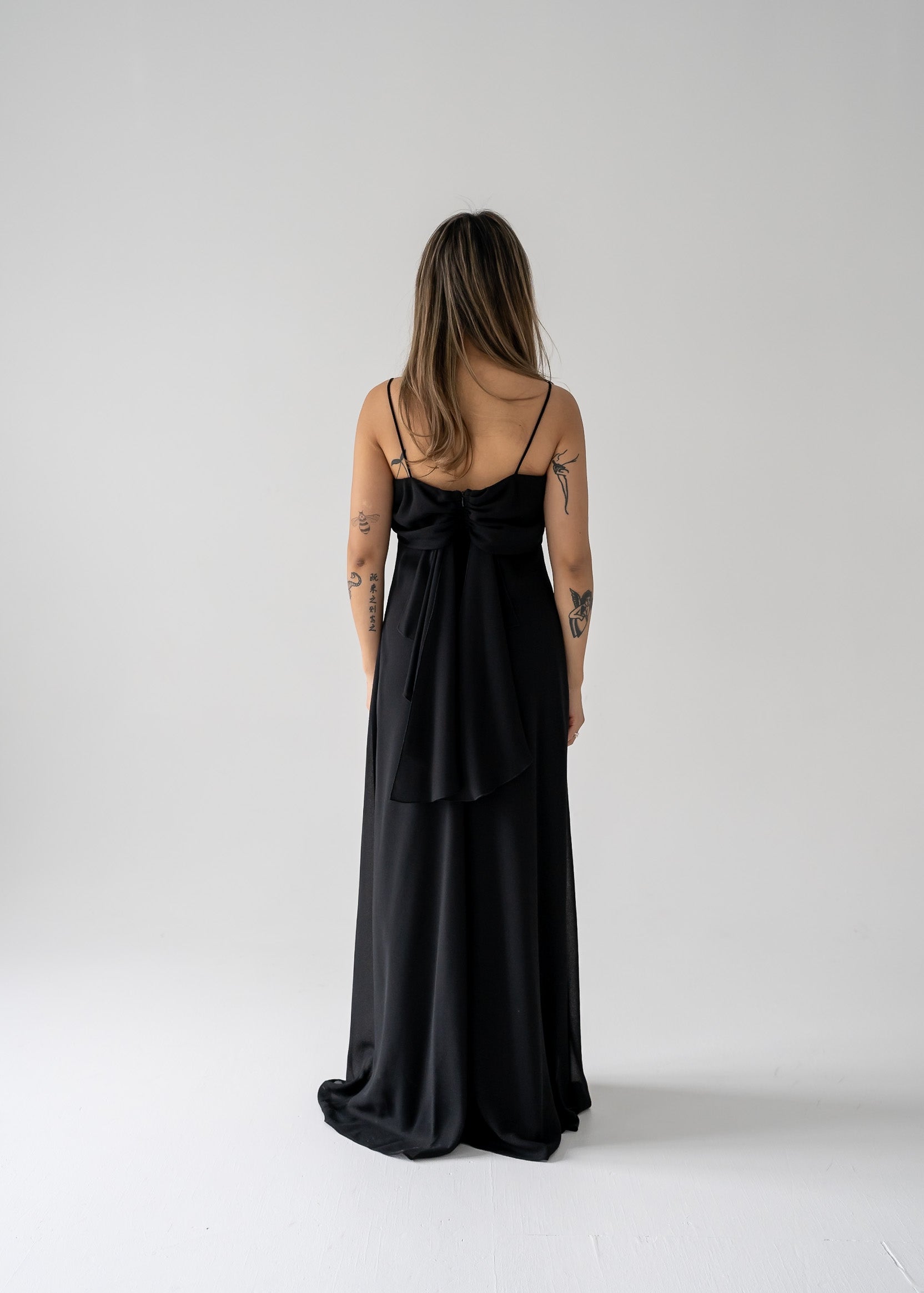 A woman standing in a studio setting, wearing a black floor-length crepe evening dress with a triple draped cowl collar and thin straps.