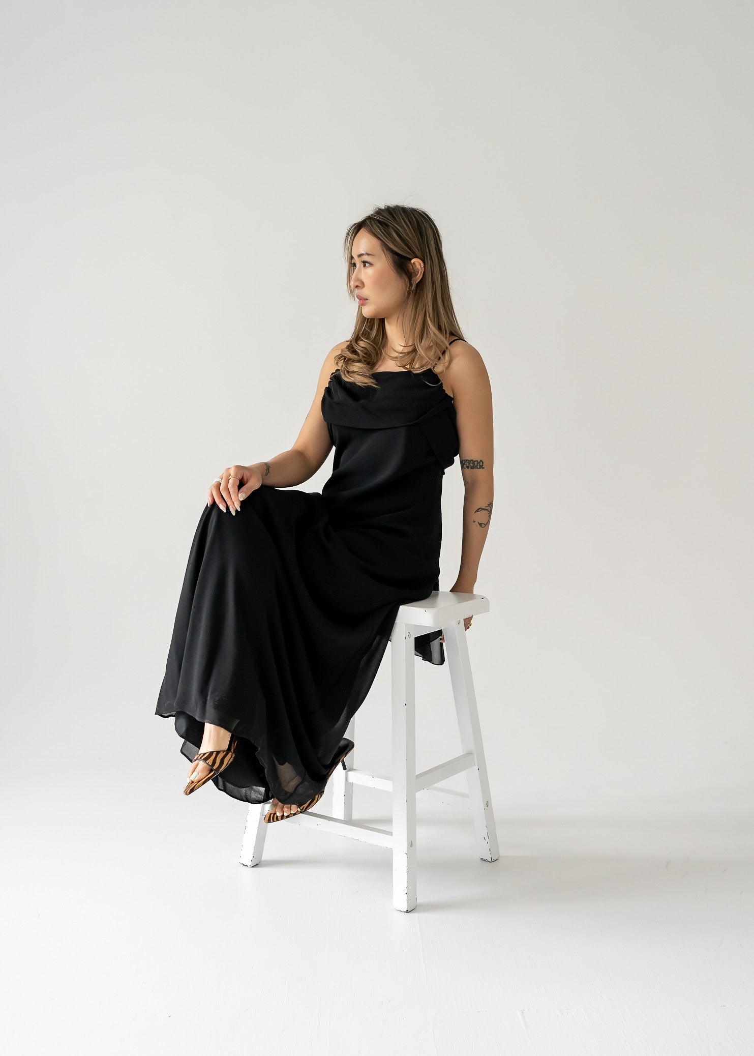 Woman in a floor length vintage black crepe dress sitting on a white stool against a plain background