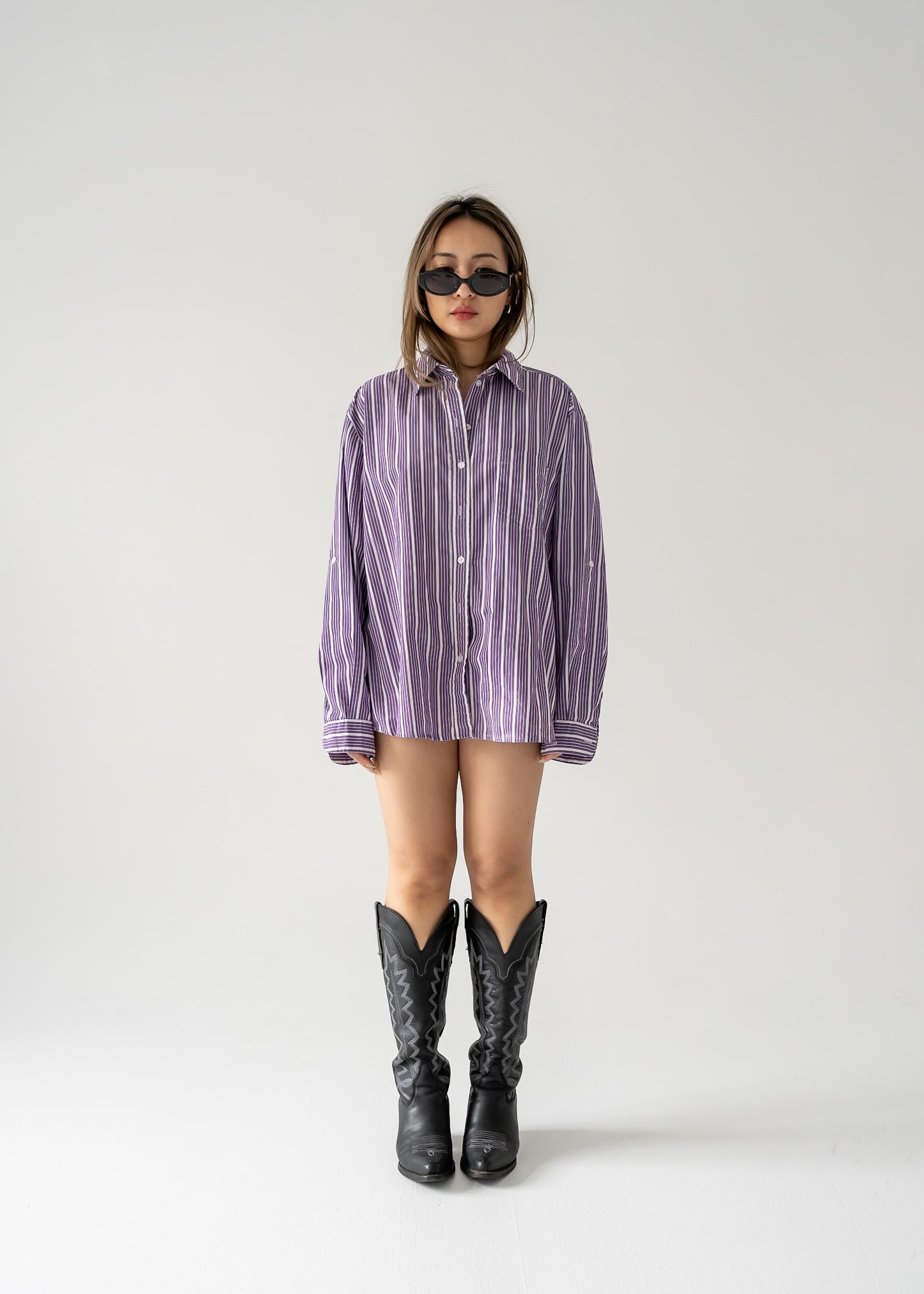 A person standing against a white background wearing a vintage purple and white striped long sleeve button-up shirt and black boots.