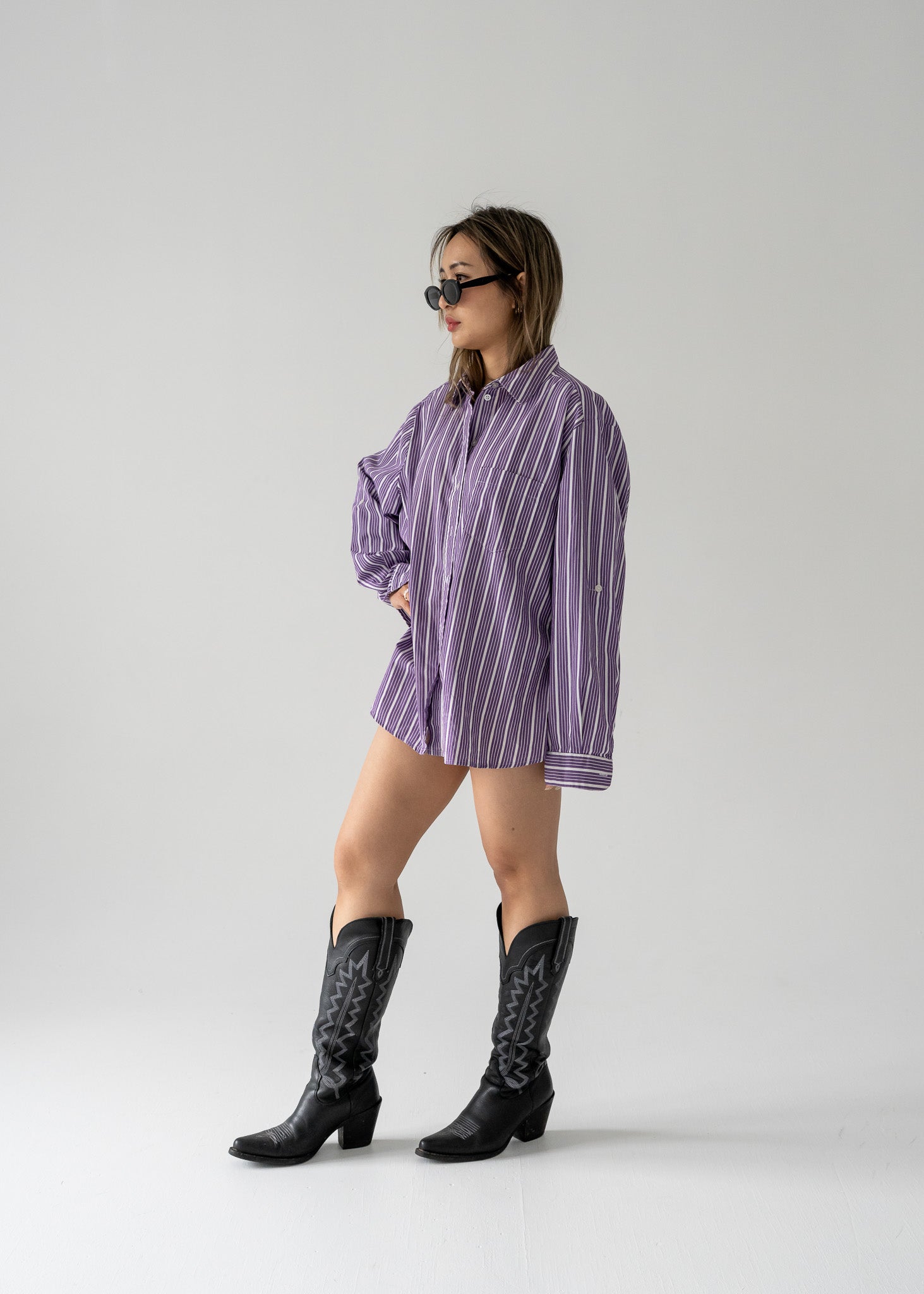 A person standing against a white background wearing a vintage purple and white striped long sleeve button-up shirt and black boots.
