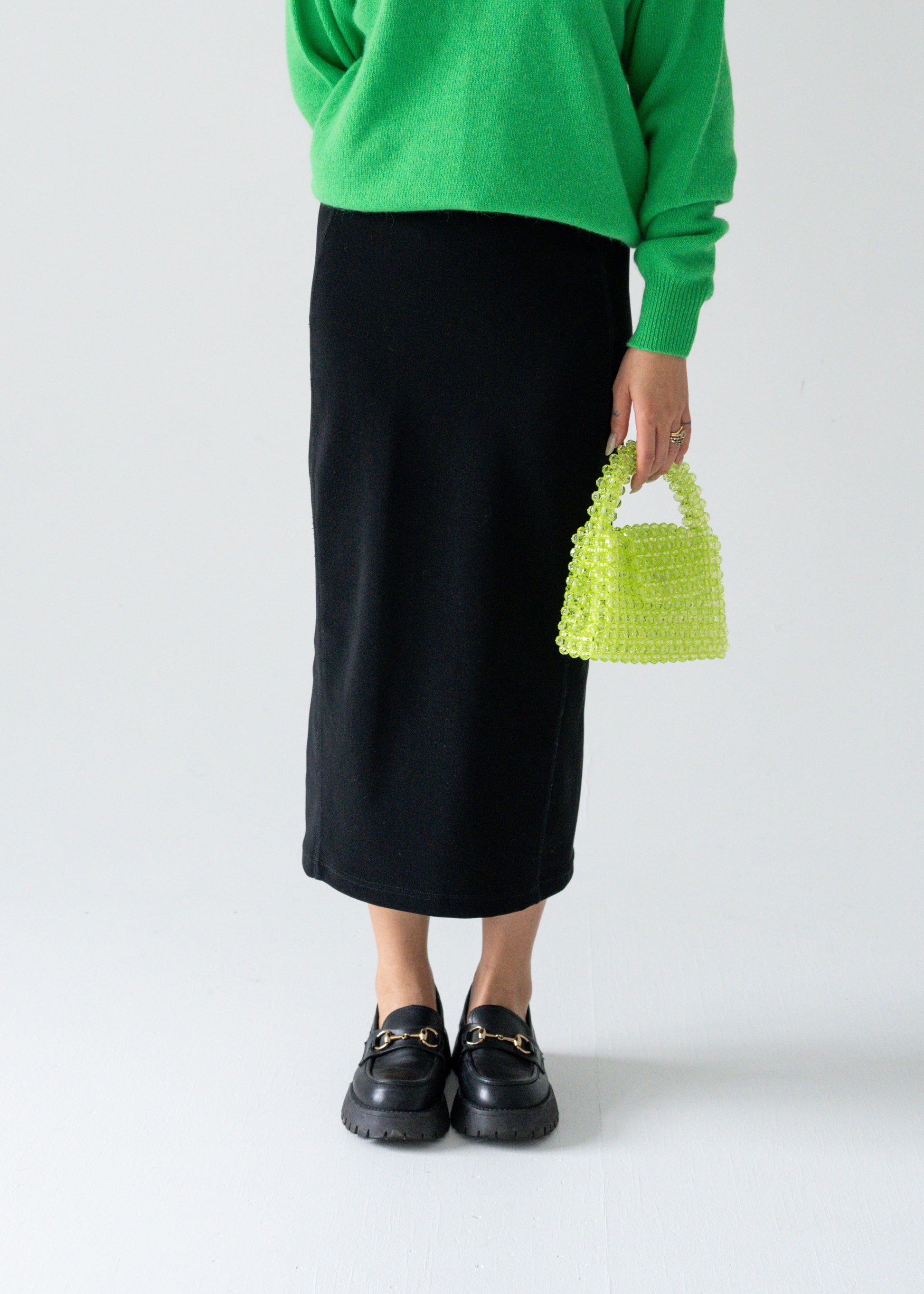 A person wearing a green sweater and a vintage black column midi skirt with a lime green handbag and black shoes.