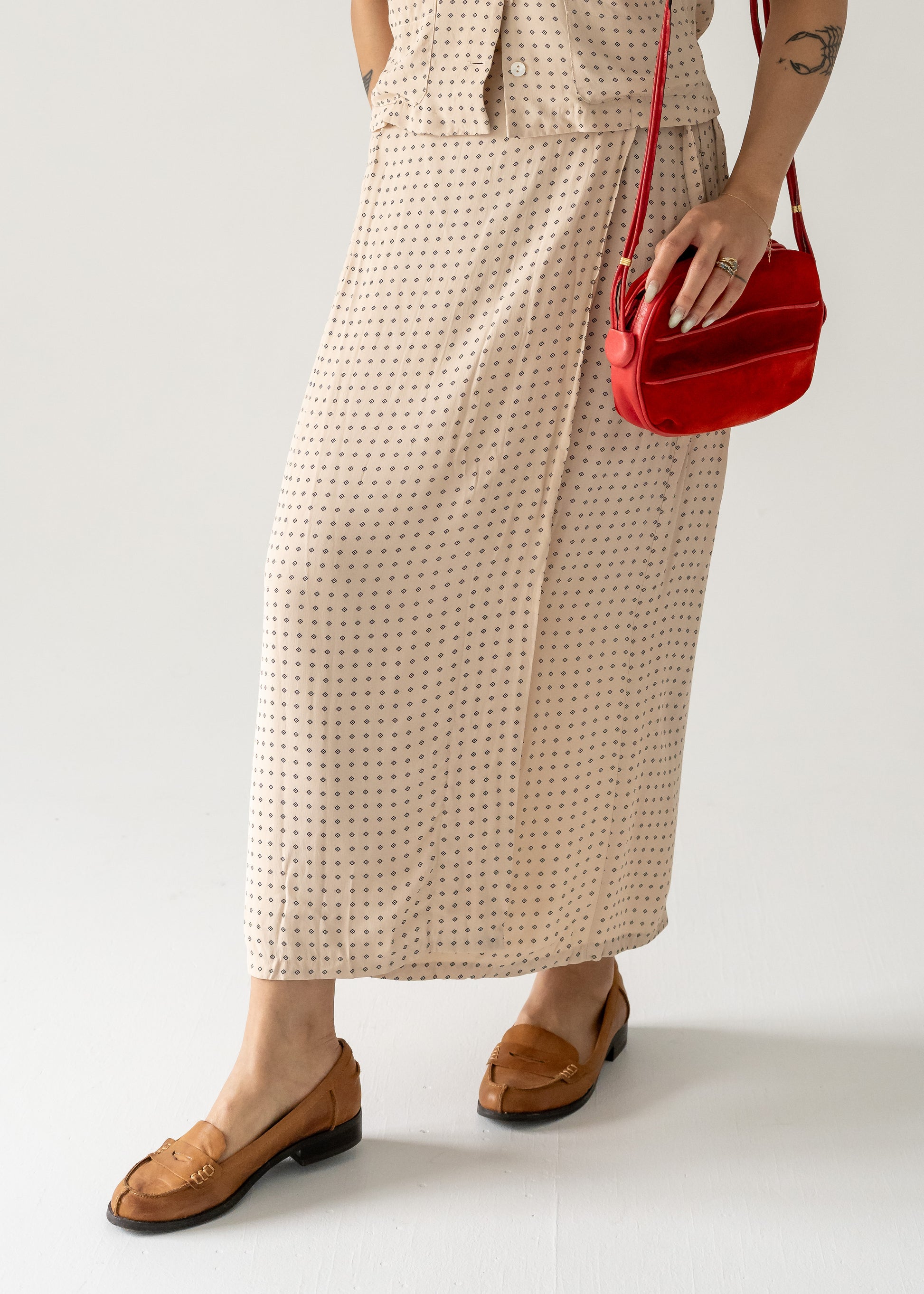 A person wearing a dotted dress and tan shoes, holding a vintage strawberry red suede leather crossbody bag with small gold hardware.