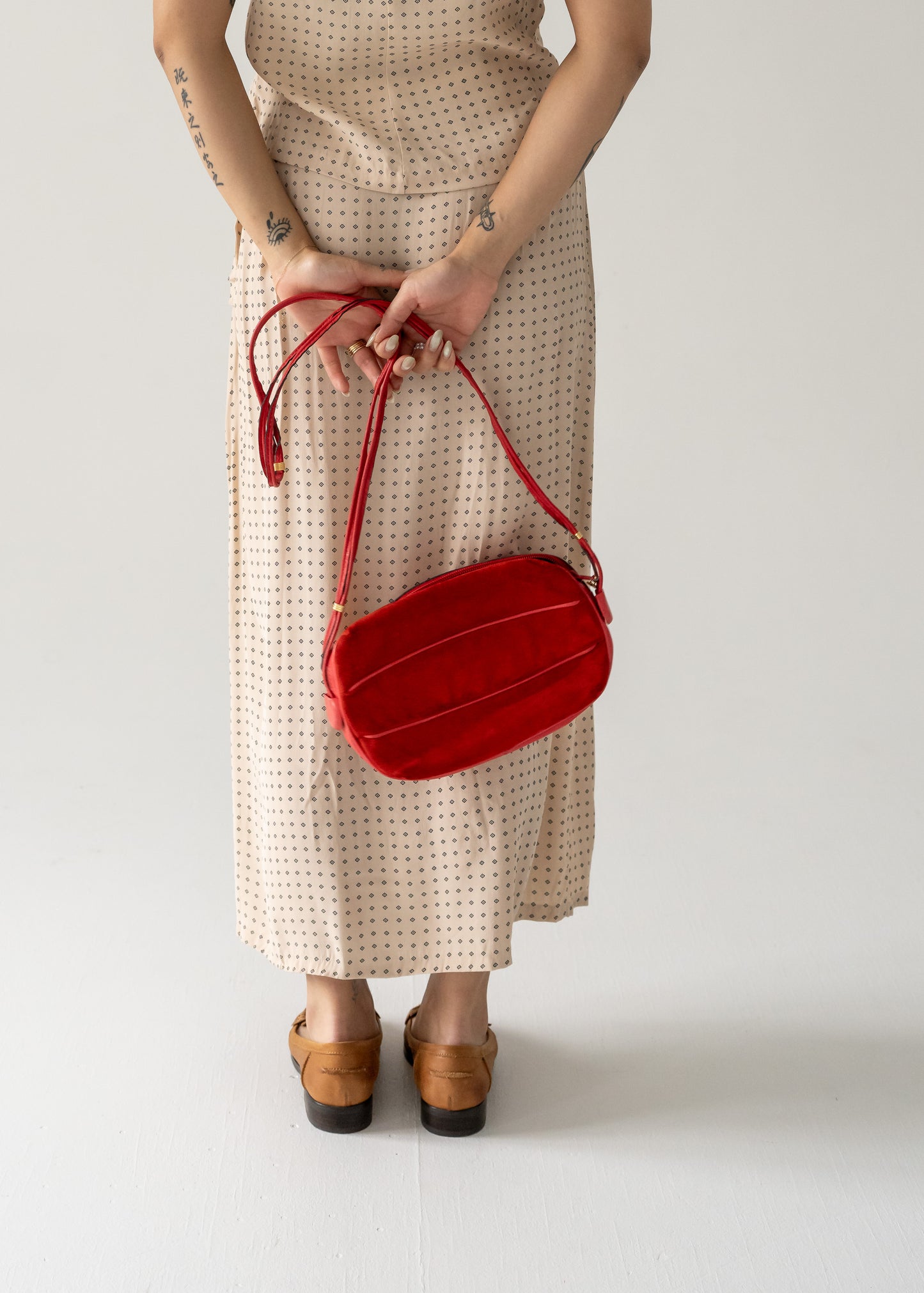 A person wearing a dotted dress and tan shoes, holding a vintage strawberry red suede leather crossbody bag with small gold hardware.
