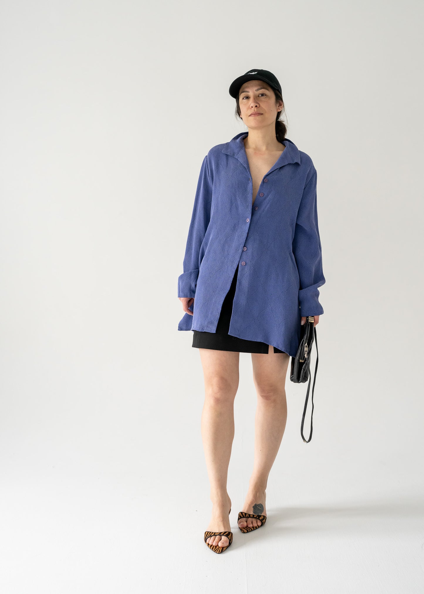 A person standing in a studio setting, wearing a vintage violet collared button front blouse with black shorts and accessories.