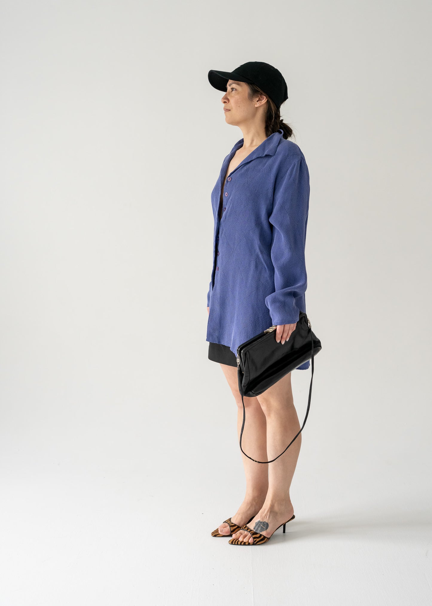 Woman in vintage violet collared button front blouse and black hat holding a black handbag against a plain background.