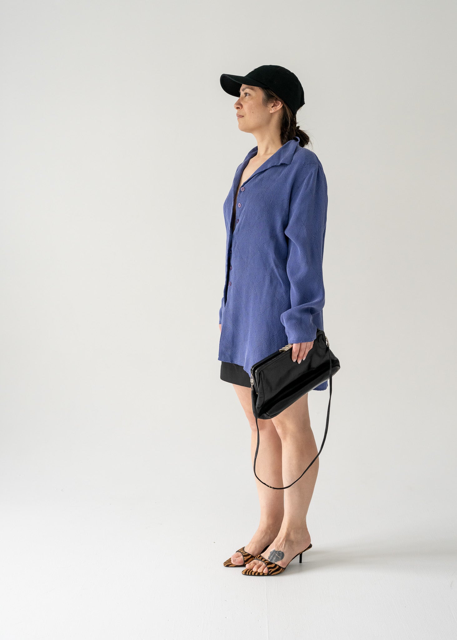 Woman in vintage violet collared button front blouse and black hat holding a black handbag against a plain background.