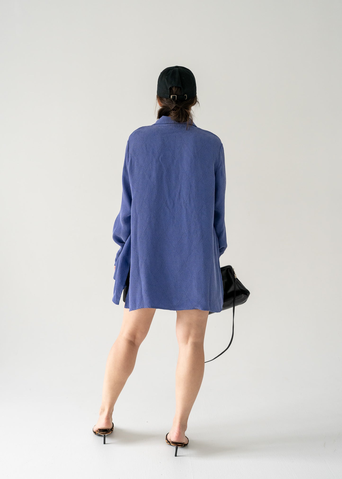 Person wearing a long vintage violet collared button front blouse with a black bag on a white background