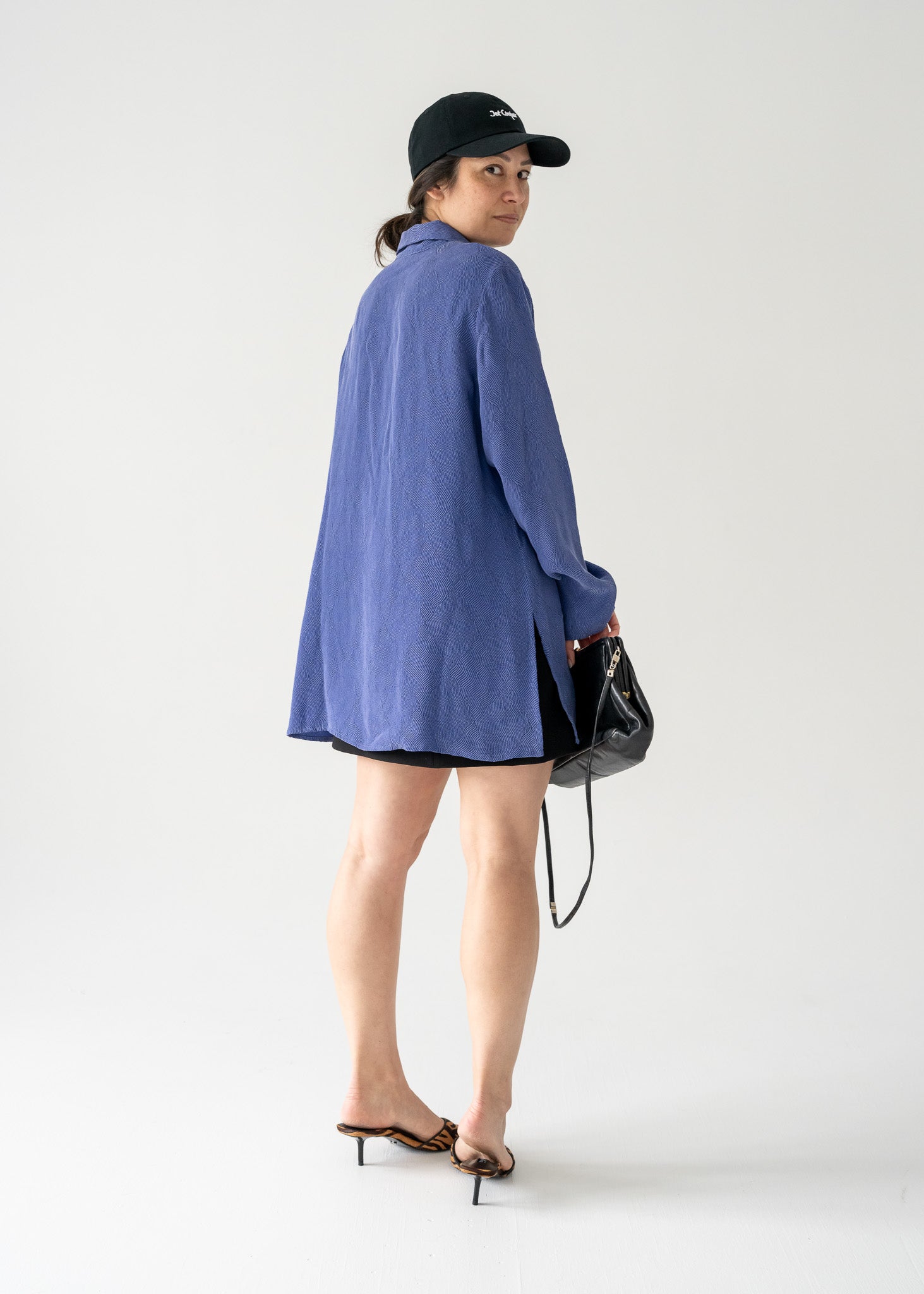 Person wearing a vintage violet collared button front blouse and black cap holding a black bag.