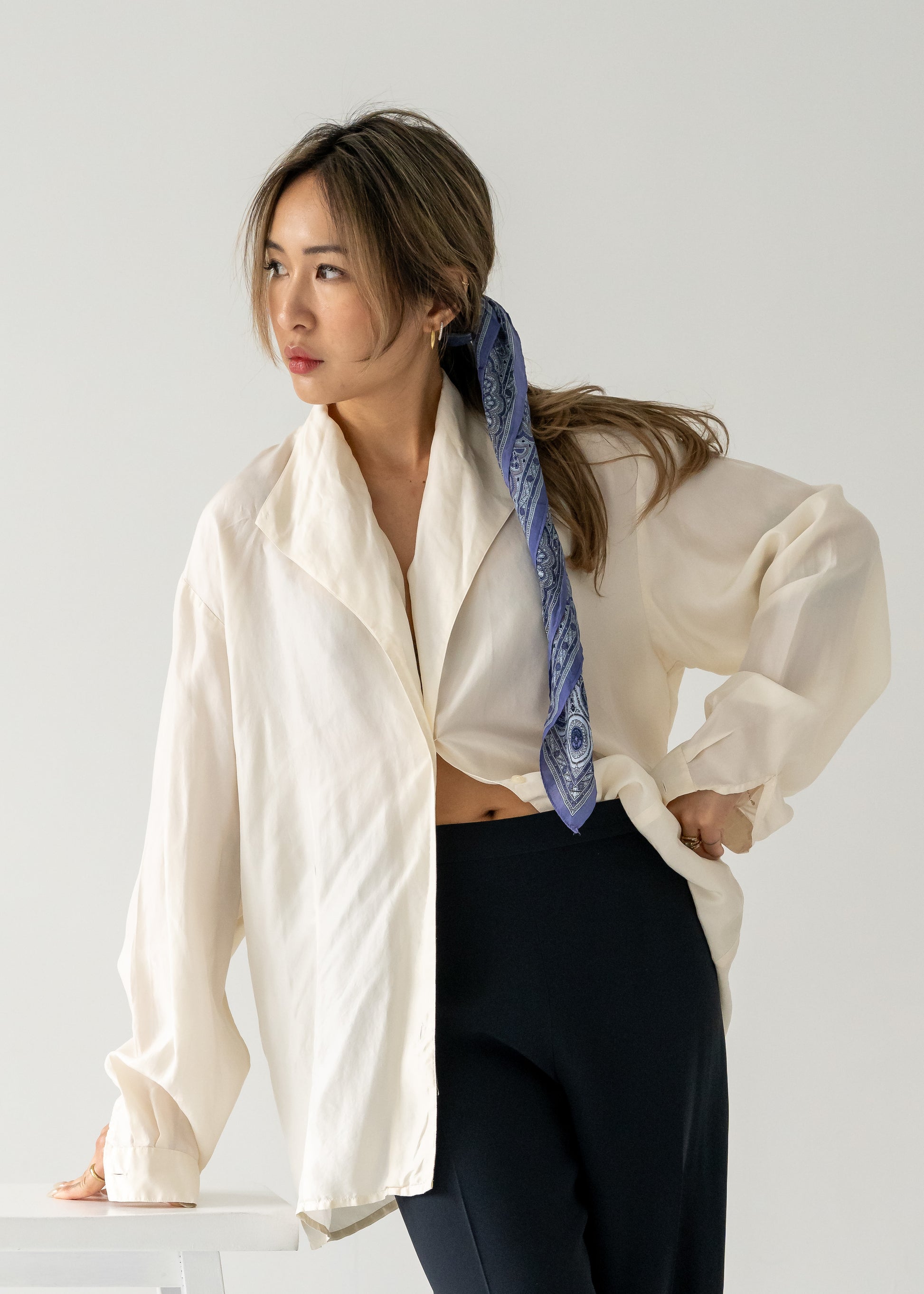 Woman wearing a white shirt and dark pants with a purple and blue silk scarf tied in her hair, standing against a plain background.