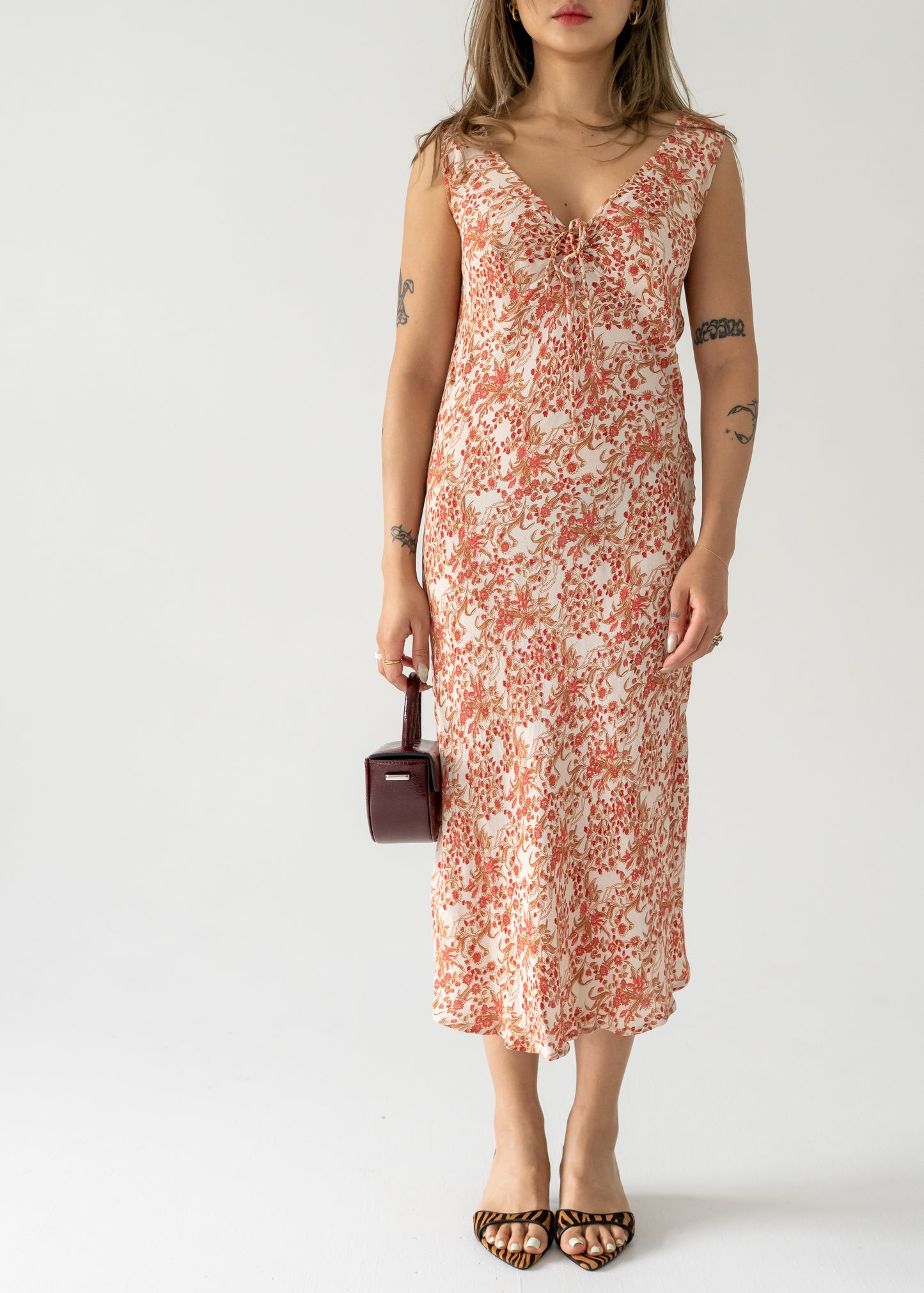 A model wearing a vintage sleeveless, cream and pink floral print dress with a cinch top and bias cut, paired with animal patterned shoes and a brown purse.